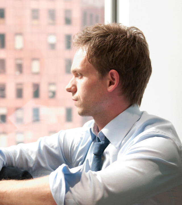 "Suits - Überholmanöver": Mike (Patrick J. Adams) sitzt in einem Hemd mit hochgekrempelten Ärmeln vor einem großen Fenster. Er blickt nachdenklich in die Ferne, das Licht fällt hell auf sein Gesicht, im Hintergrund ist die Fassade eines Hochhauses zu sehen.