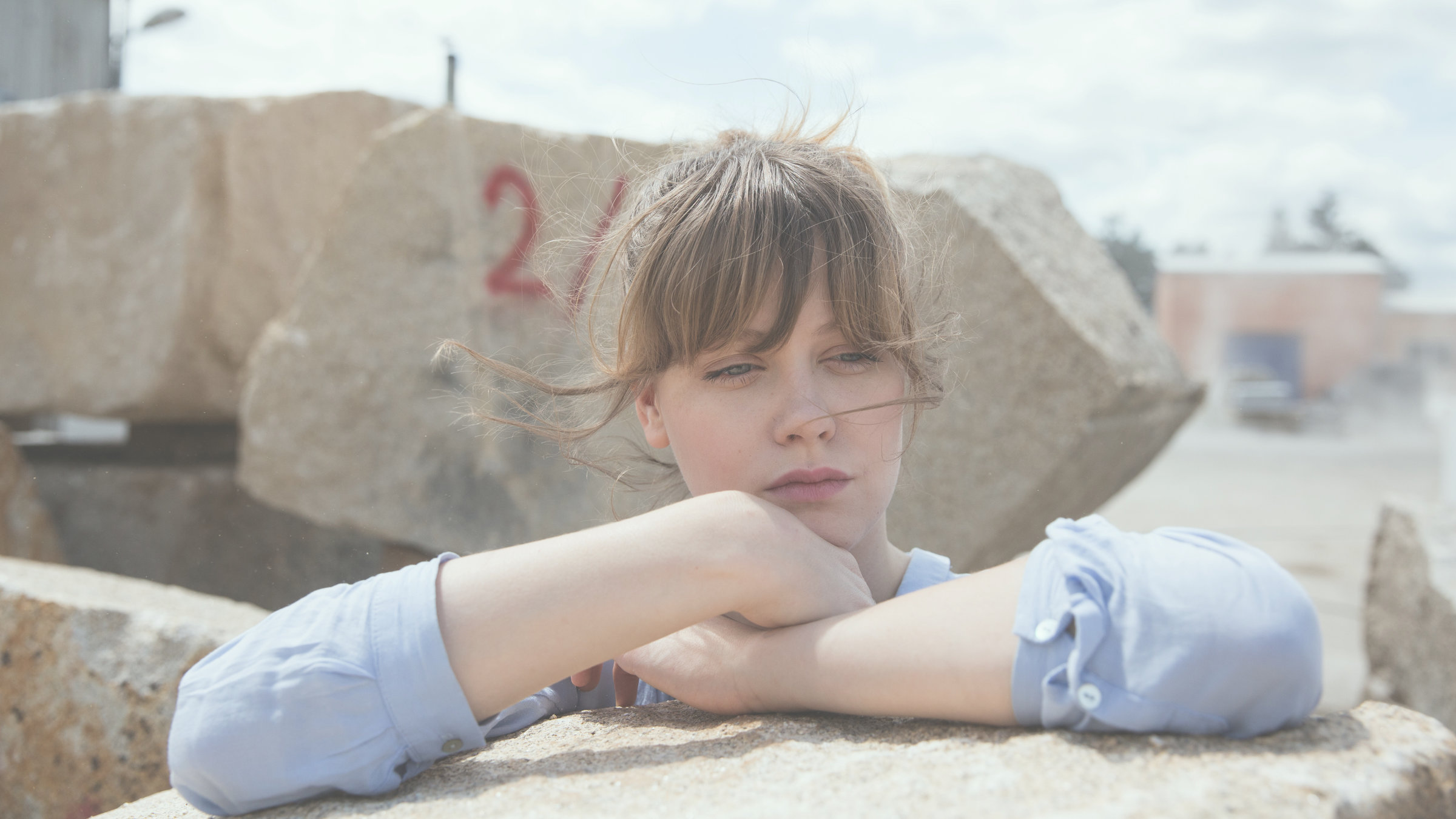"Momente": Eine junge Frau mit zerzausten halblangen dunkelblonden Haaren schaut, die Oberarme auf einen hohen Stein gestützt, verträumt nach links unten aus dem Bild. Hinter ihr steht eine Art Mauer weiterer grob zugeschnittener unregelmäßiger Steinklötze; einer ist mit einer roten „2“ gekennzeichnet.
