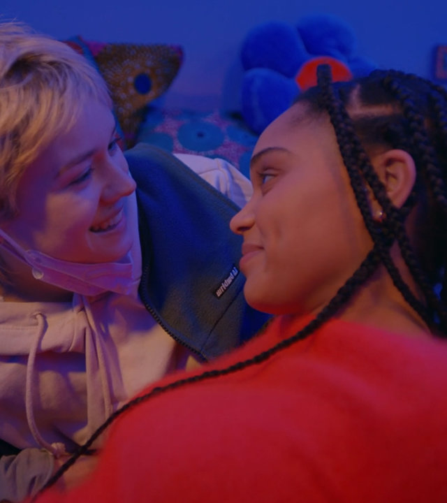 "Becoming Charlie - Fehl am Platz": Charlie (Lea Drinda) und Ronja (Sira Anna Faal) liegen sich auf einem Bett gegenüber und sehen sich an. Das Zimmer ist blau beleuchtet. Im Hintergrund sind bunte Kissen zu sahen sowie ein Plakat an der Wand.