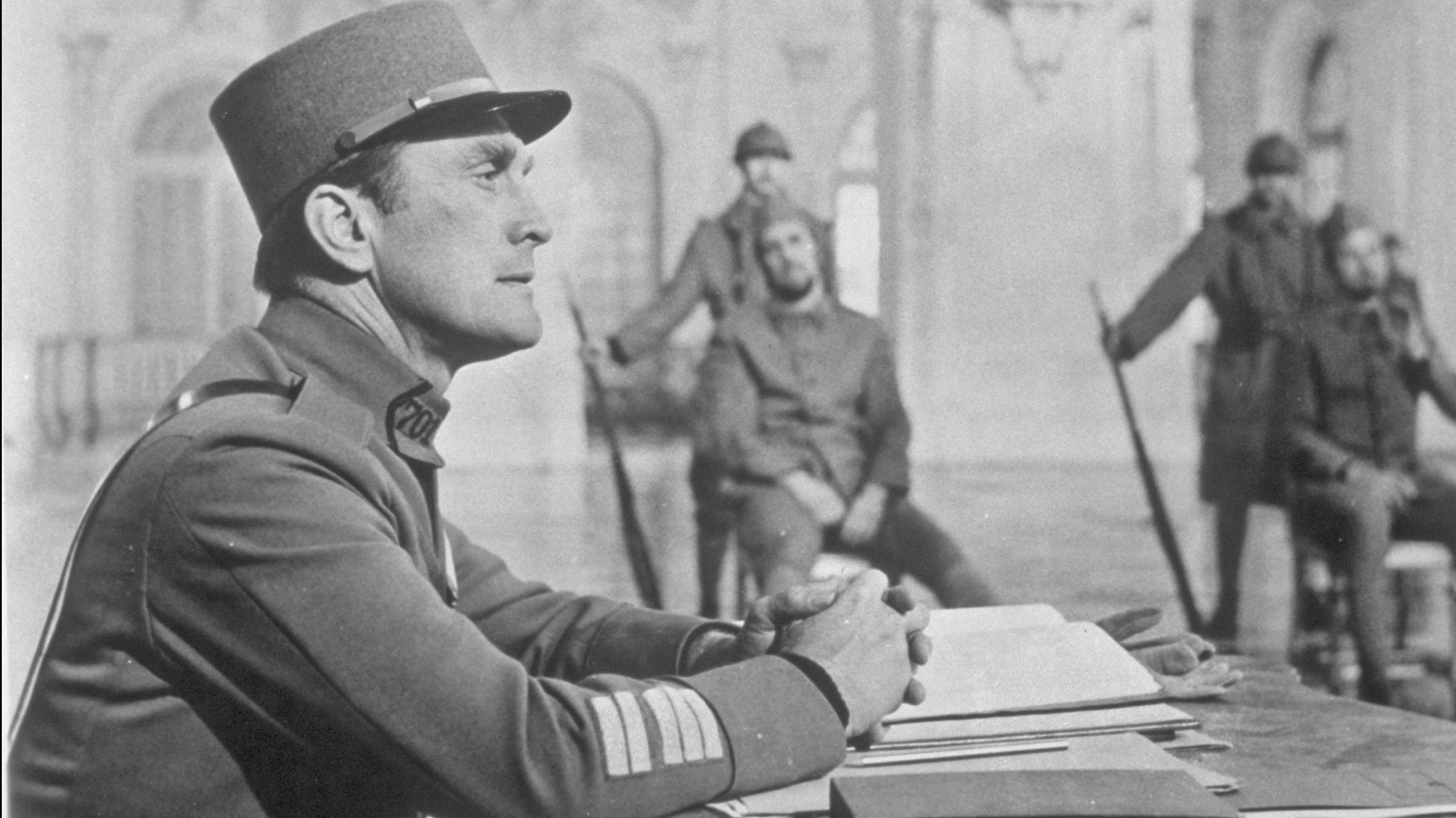 "Wege zum Ruhm": Ein Mann in Uniform (Kirk Douglas) sitzt links im Bildvordergrund und blickt konzentriert nach vorne. Im Bildhintergrund sieht man leicht unscharf zwei angeklagte Männer, hinter denen jeweils eine Wache mit Gewehr steht.