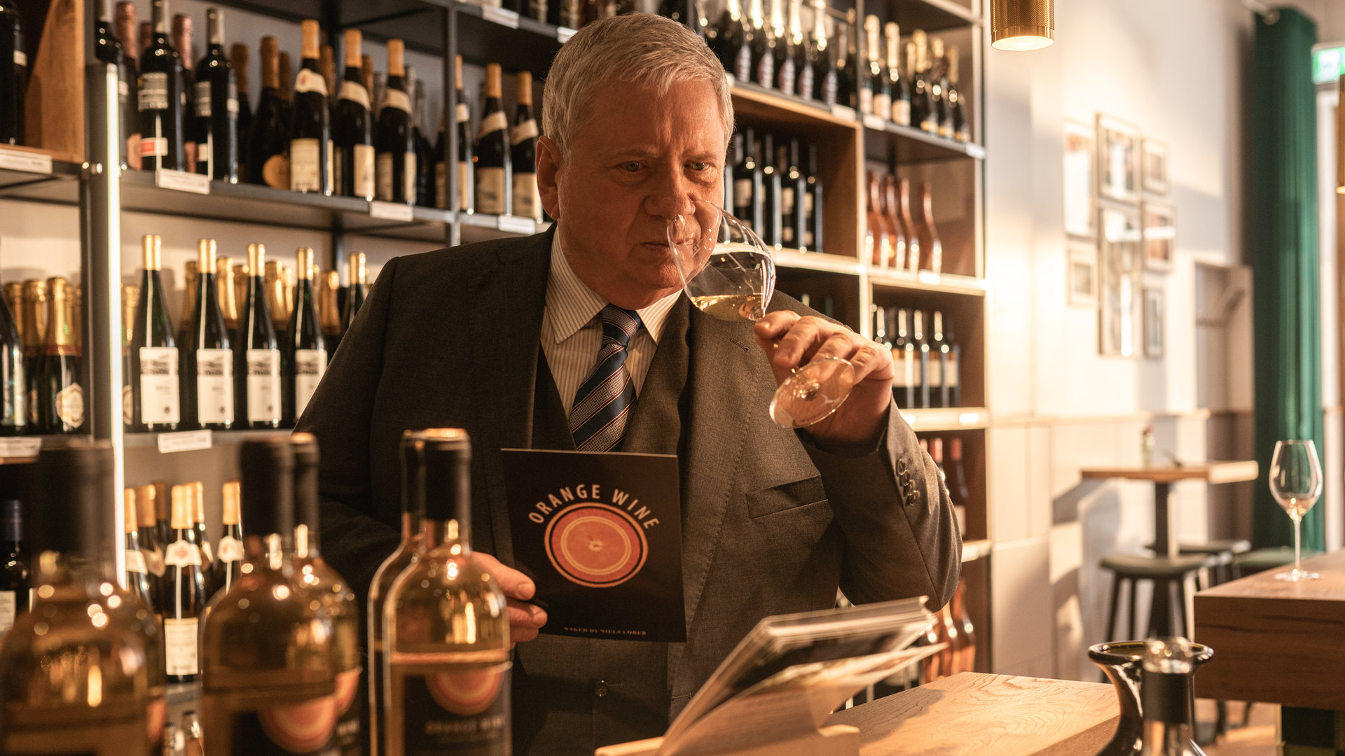 "Der Staatsanwalt - Wein und Mord": Oberstaatsanwalt Bernd Reuther (Rainer Hunold) steht an der Theke einer Weinhandlung. In der linken Hand hält er ein Weinglas mit einem Schluck Wein, an dem er vorsichtig riecht. In der rechten Hand hält er einen Flyer über Weine.
