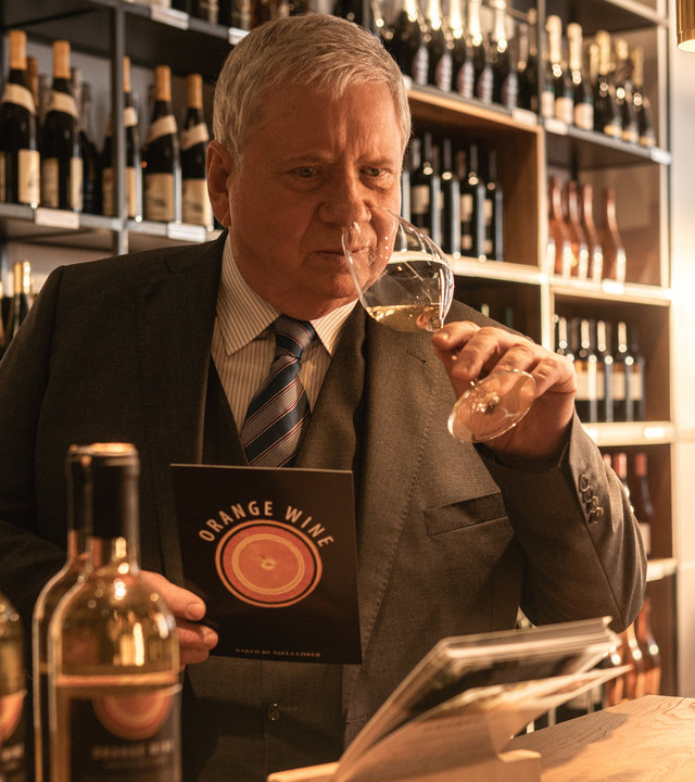 "Der Staatsanwalt - Wein und Mord": Oberstaatsanwalt Bernd Reuther (Rainer Hunold) steht an der Theke einer Weinhandlung. In der linken Hand hält er ein Weinglas mit einem Schluck Wein, an dem er vorsichtig riecht. In der rechten Hand hält er einen Flyer über Weine.
