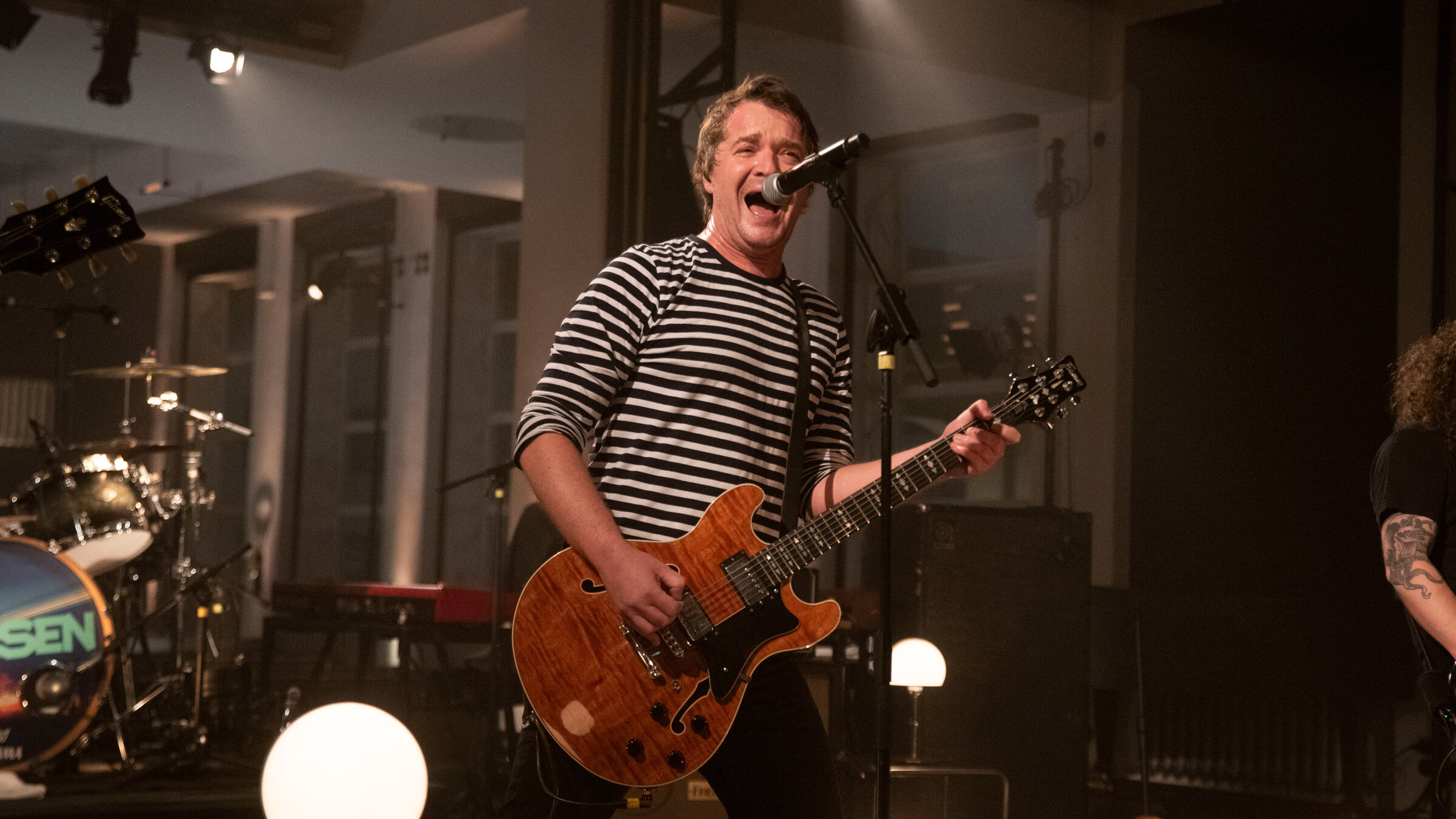 "Pop Around@Bauhaus: Madsen": Der Sänger Madsen steht auf der Bühne und singt. Er begleitet sich dabei auf einer Gitarre. Vor ihm steht ein Mikrofonständer. Gekleidet ist er mit einem schwarz-weiß gestreiften Pullover und einer schwarzen Hose. Links im Anschnitt ist ein Teil eines Schlagzeugs zu erkennen.