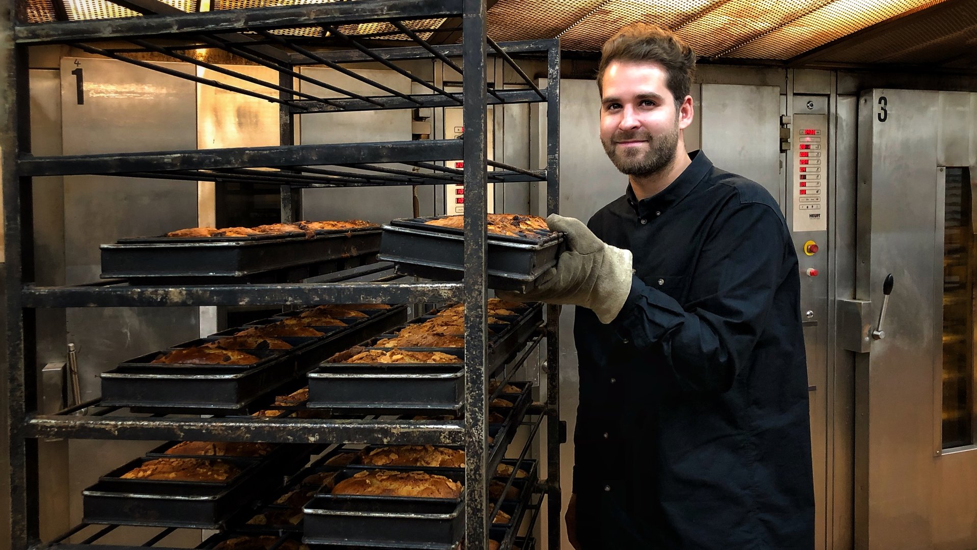 "Terra Xpress - Mit gutem Gewissen - guten Appetit!": Der Bäcker Lars Peters steht in der Backstube und schaut lächelnd in die Kamera. Er steht neben einem Rollwagen und hält ein Blech mit Bananenbroten in der linken Hand, welches wie fünf andere gerade frisch aus dem Ofen kommt.