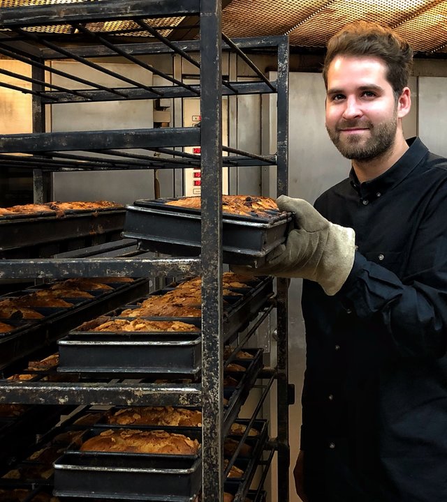 "Terra Xpress - Mit gutem Gewissen - guten Appetit!": Der Bäcker Lars Peters steht in der Backstube und schaut lächelnd in die Kamera. Er steht neben einem Rollwagen und hält ein Blech mit Bananenbroten in der linken Hand, welches wie fünf andere gerade frisch aus dem Ofen kommt.