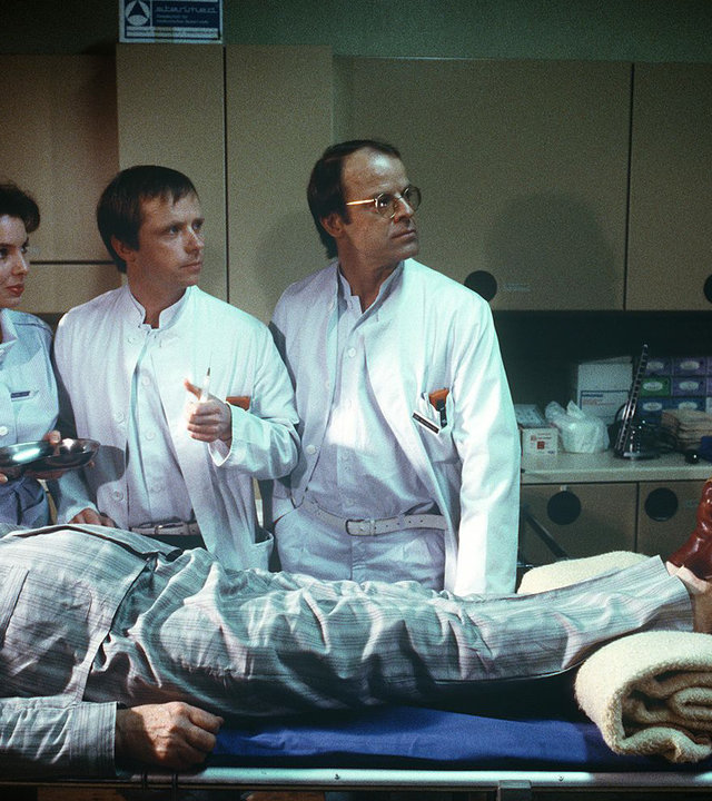 "Die Schwarzwaldklinik - Der zudringliche Patient": Schwester Ina Gaby Fischer), Dr. Engel Michael Kausch) und Dr. Borsdorf Knut Hinz) stehen an einer Behandlungsliege auf der ein bewusstloser Patient (Darsteller unbekannt) liegt und sehen zu Herrn Körtler (Klaus Guth), der am Fußende steht.