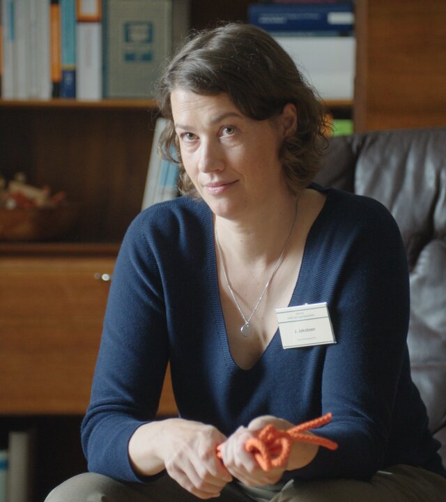 "Hungry - Crush": Therapeutin Frau Jakobsen (Minna Wündrich) sitzt in ihrem Büro auf einem braunen Ledersessel vor einer mit Fachliteratur gefüllten Schrankwand. Sie hält eine orangefarbene Kordel in der Hand und schaut zur Seite.