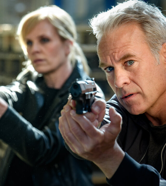 "SOKO Leipzig - Was bleibt (1)": Ina Zimmermann (Melanie Marschke) und Jan Maybach (Marco Girnth) stehen mit gezogenen Waffen hinter einem Stapel Paletten.