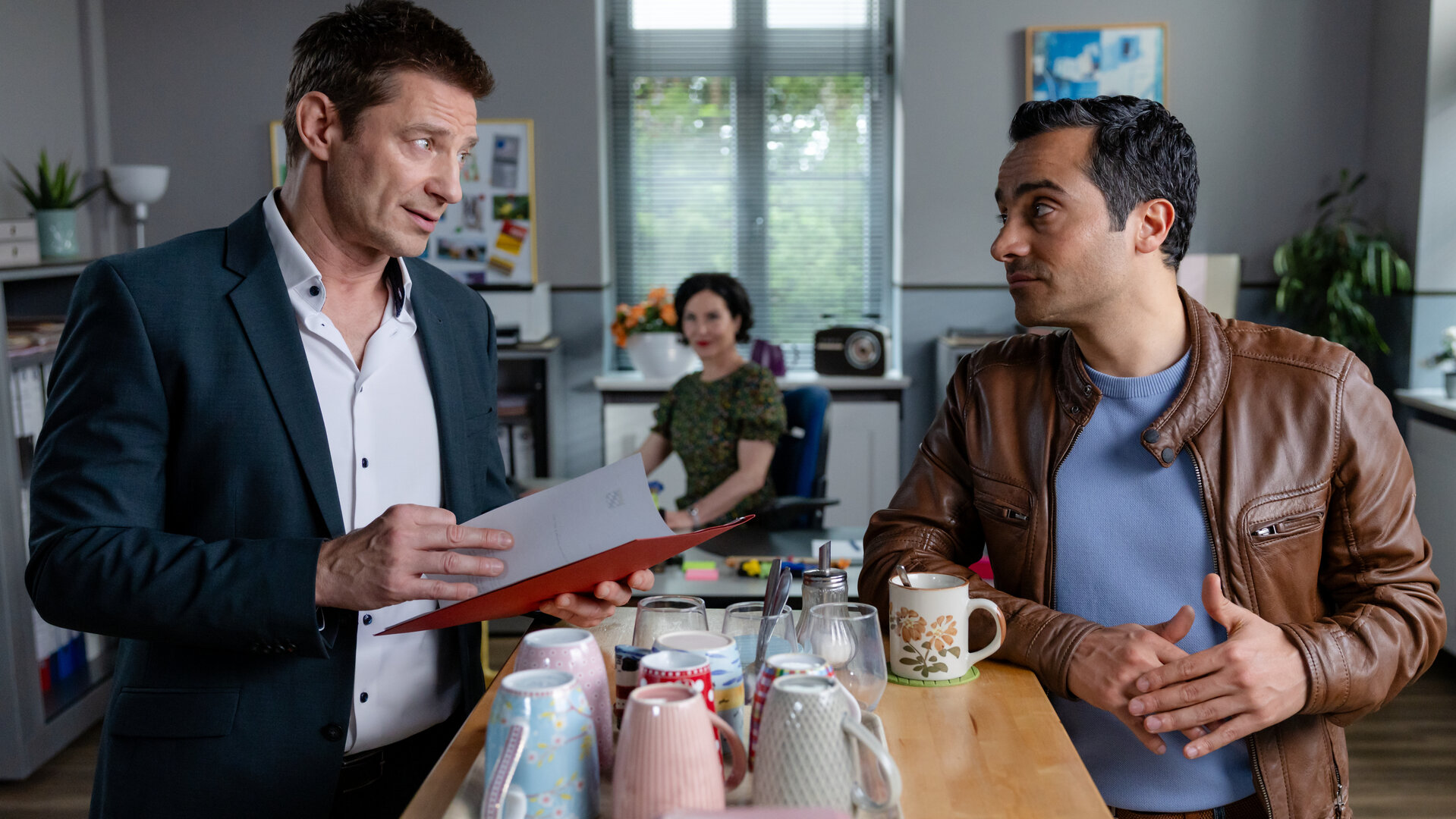 "Die Rosenheim-Cops - Ein klarer Fall": Hauptkommissar Hansen (Igor Jeftić,) und Kommissar Kaya (Baran Hêvî) stehen in Frau Stockls (Marisa Burger) Büro am Kaffeetresen und unterhalten sich. Hansen hält eine Akte in den Händen, während er Kaya ansieht. Auf dem Tresen steht ein Tablett mit Kaffeebechern. Im Hintergrund sitzt Frau Stockl am Schreibtisch und schaut zu den beiden.