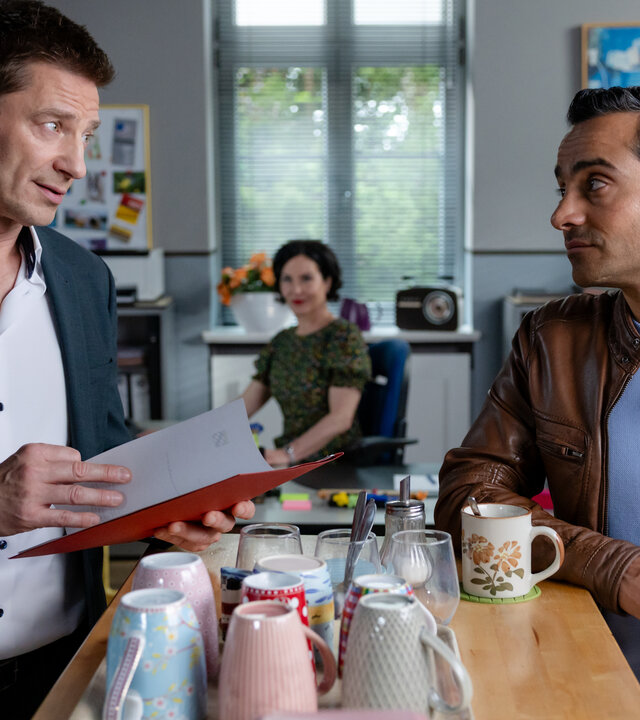 "Die Rosenheim-Cops - Ein klarer Fall": Hauptkommissar Hansen (Igor Jeftić,) und Kommissar Kaya (Baran Hêvî) stehen in Frau Stockls (Marisa Burger) Büro am Kaffeetresen und unterhalten sich. Hansen hält eine Akte in den Händen, während er Kaya ansieht. Auf dem Tresen steht ein Tablett mit Kaffeebechern. Im Hintergrund sitzt Frau Stockl am Schreibtisch und schaut zu den beiden.