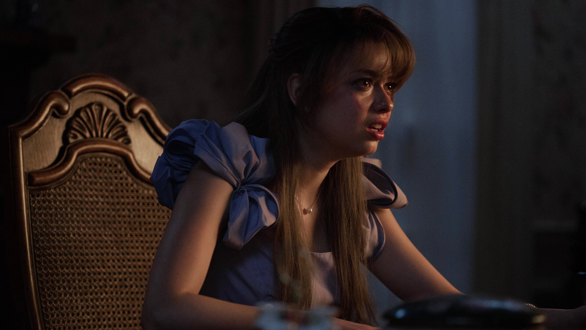 "Hysteria! - Deprogrammierung": Faith (Nikki Hahn) sitzt an einem dunklen Esstisch und blickt mit schockiertem Gesichtsausdruck geradeaus. Auf ihrer Wange ist eine rote Strieme zu erkennen.