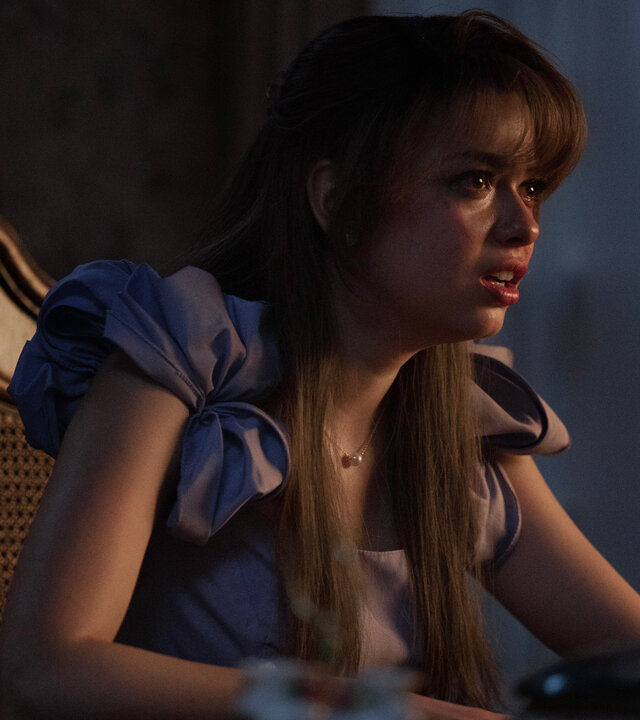 "Hysteria! - Deprogrammierung": Faith (Nikki Hahn) sitzt an einem dunklen Esstisch und blickt mit schockiertem Gesichtsausdruck geradeaus. Auf ihrer Wange ist eine rote Strieme zu erkennen.