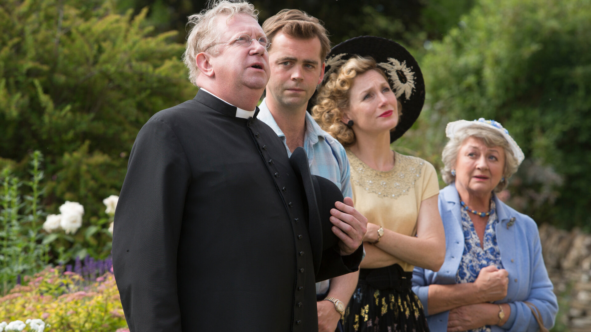 "Father Brown - Die Moral des Henkers": Father Brown (Mark Williams), Sid Carter (Alex Price), Lady Felicia (Nancy Carroll) und Mrs. McCarthy (Sorcha Cusack) stehen nebeneinander im Garten und schauen in eine Richtung.