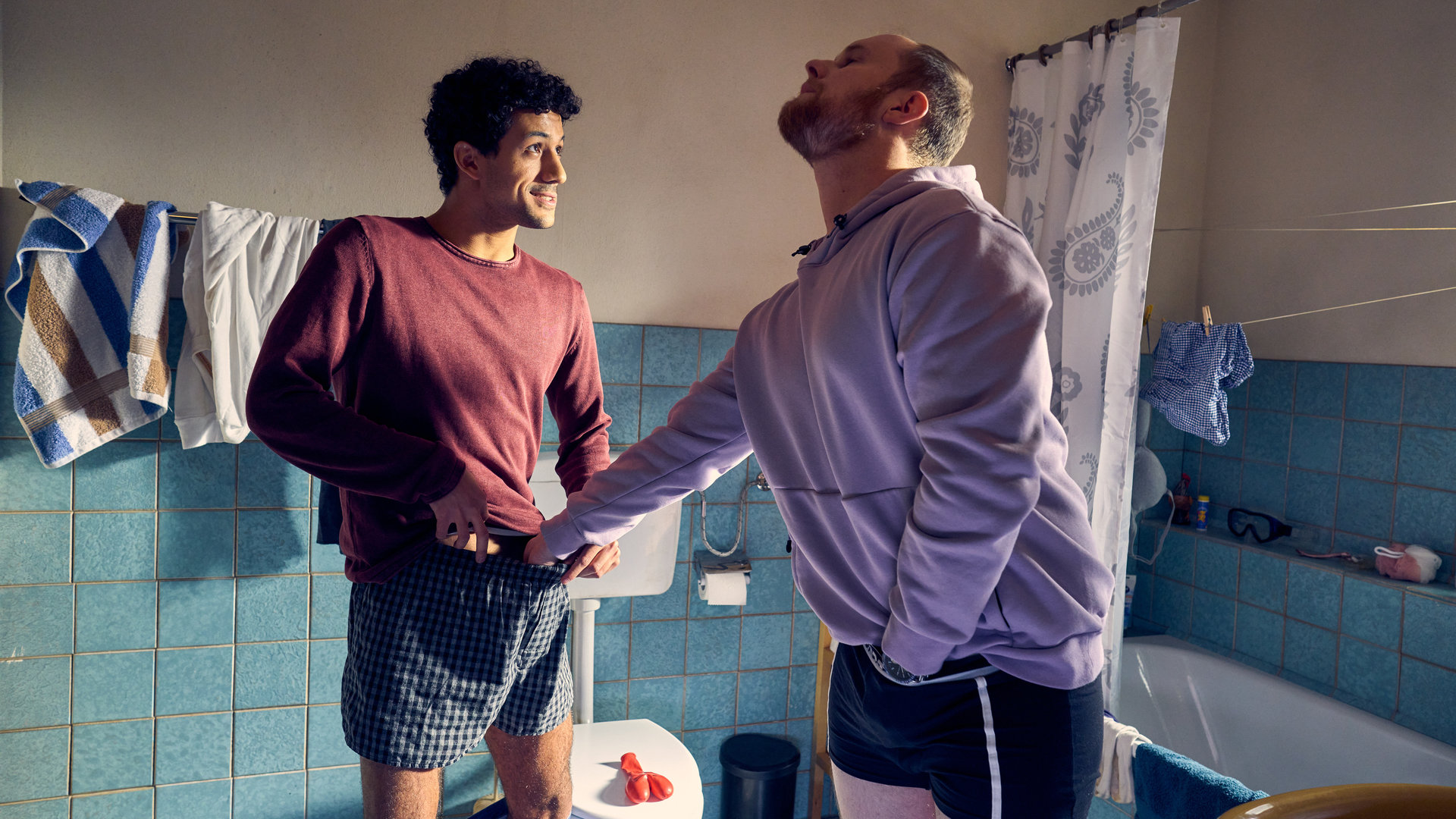 "Ich dich auch! - Der Knubbel": Yannik (Rojan Juan Barani) und Sven (Sven Daniel Bühler) sind im Badezimmer. Sven fasst sich mit einer Hand in die Unterhose, mit der anderen zieht er an Yanniks Boxershorts. Yannik schaut ihn lächelnd an.