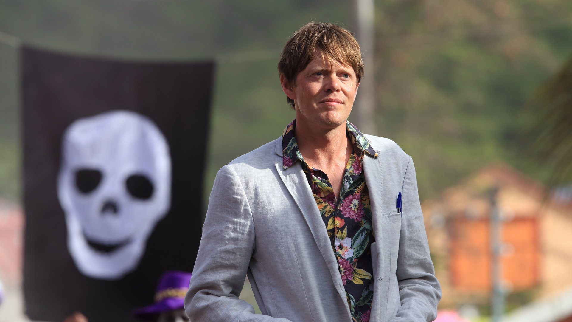 "Death in Paradise: Der Mann, den es nicht gab": Humphrey (Kris Marshall) steht im Freien vor einer schwarzen Flagge auf der ein weißer Totenkopf zu sehen ist.