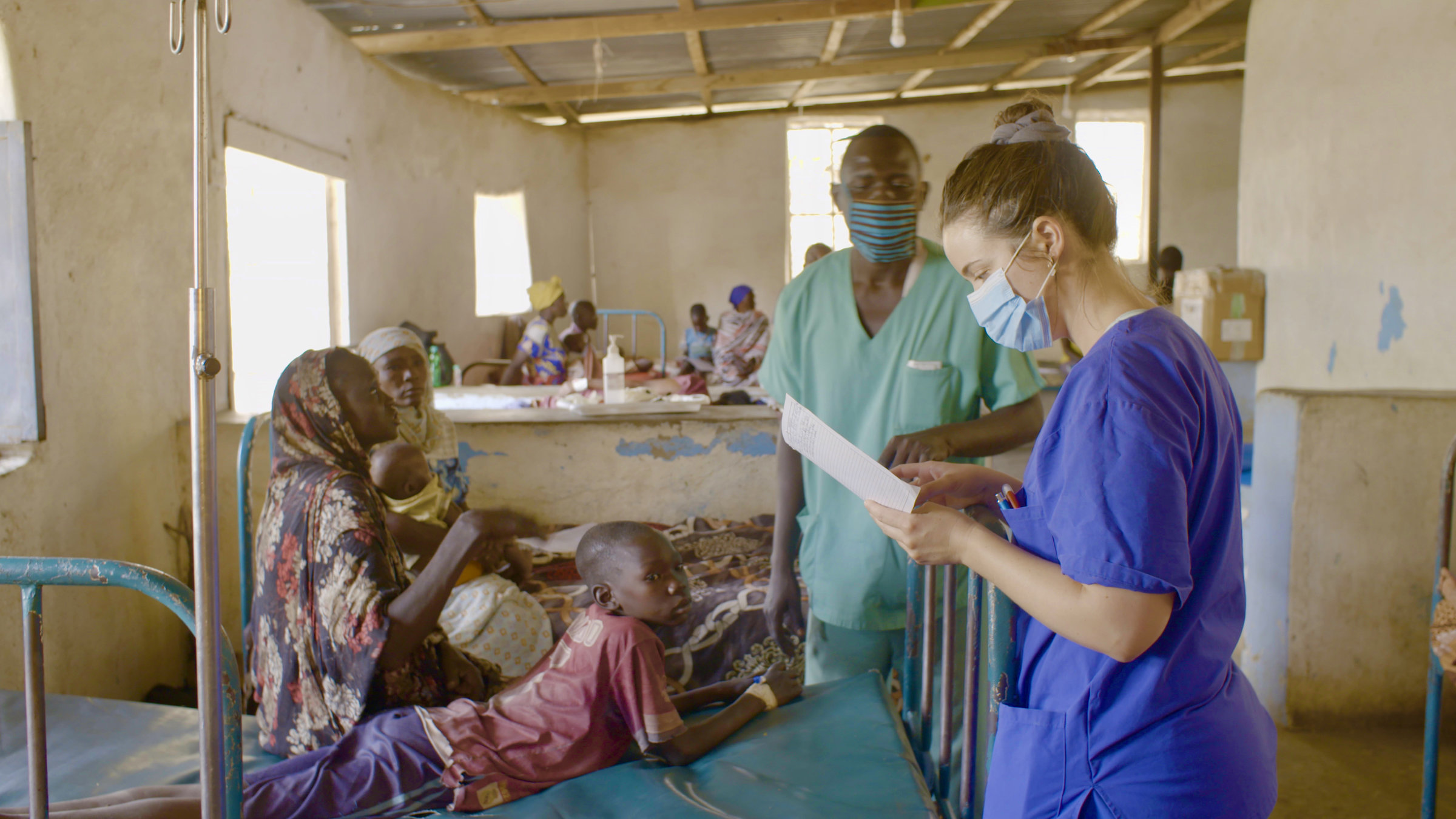 "37°: Helfen im Krisengebiet - Zwischen Grenzerfahrung und Nächstenliebe": Anke steht während ihrer Visite mit Mundschutz und in blauer Arbeitskleidung vor einem rostigen Bett, auf dem eine Sudanesin sitzt, daneben liegt ihr Sohn auf dem Bauch und schaut zur Seite. Die junge Frau spricht mit Anne. Im Hintergrund sieht man einen sudanesischen Mediziner mit Mundschutz sowie weitere Betten mit Patienten