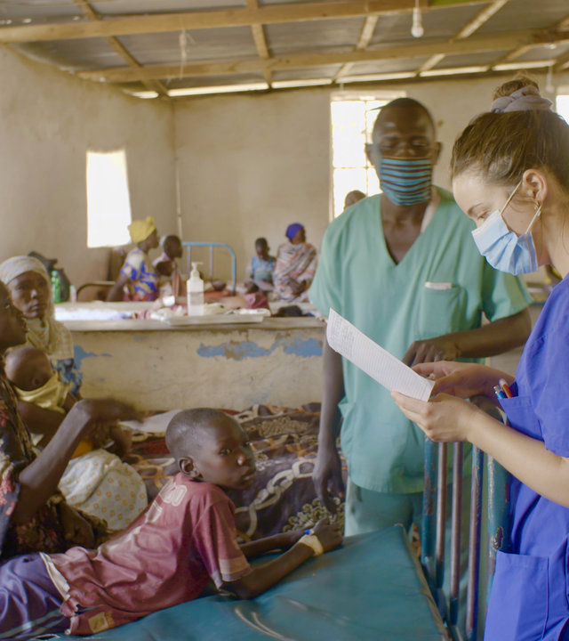 "37°: Helfen im Krisengebiet - Zwischen Grenzerfahrung und Nächstenliebe": Anke steht während ihrer Visite mit Mundschutz und in blauer Arbeitskleidung vor einem rostigen Bett, auf dem eine Sudanesin sitzt, daneben liegt ihr Sohn auf dem Bauch und schaut zur Seite. Die junge Frau spricht mit Anne. Im Hintergrund sieht man einen sudanesischen Mediziner mit Mundschutz sowie weitere Betten mit Patienten