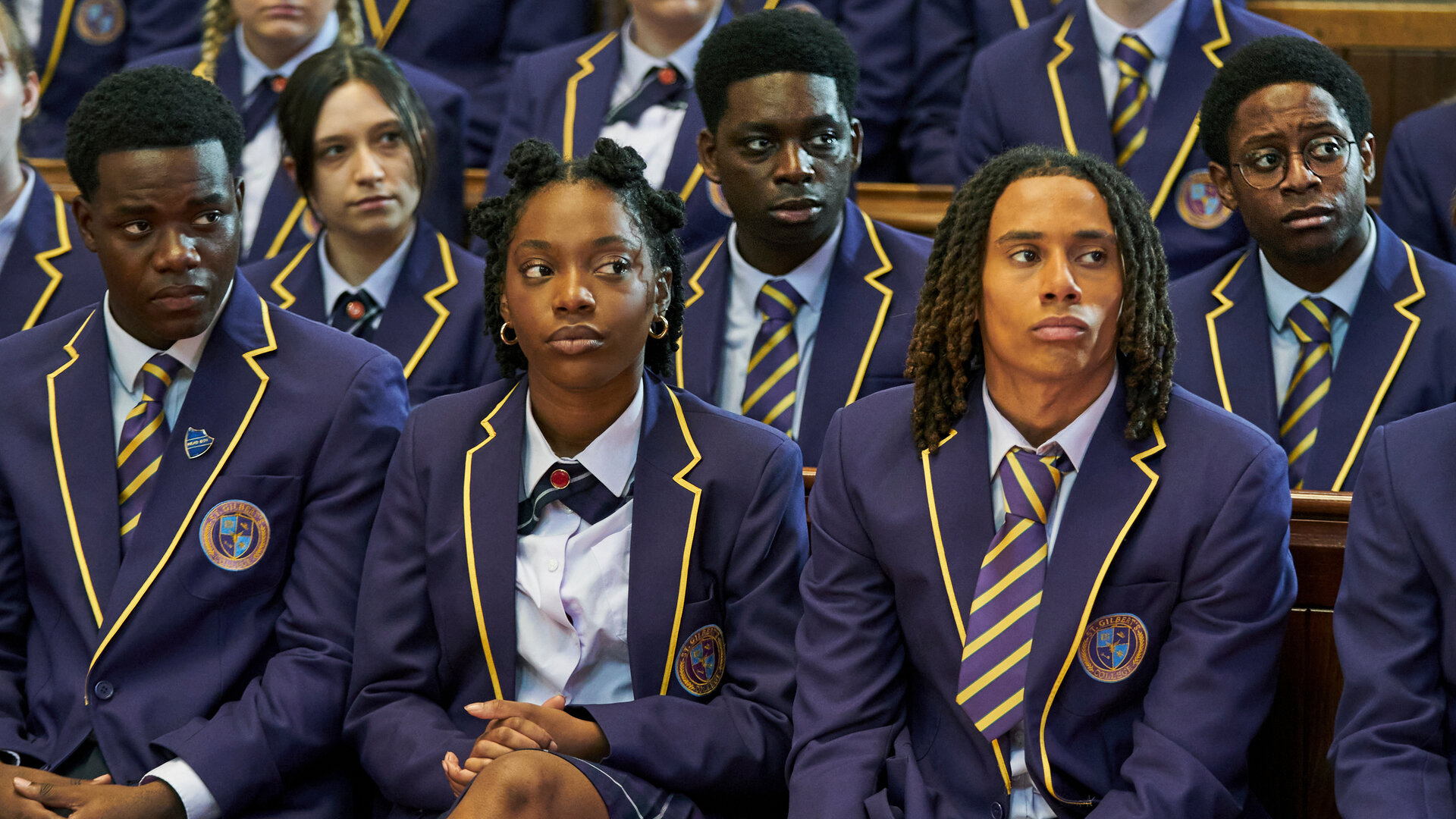 "Boarders - Welcome to St. Gilbert's- Piercings & Protests": Jaheim (Josh Tedeku), Leah (Jodie Campbell), Toby (Sekou Diaby), Omar (Myles Kamwendo) und Femi (Aruna Jalloh) sitzen mit Mitschülerinnen und Mitschülern (Komparsen) in der Aula. Alle tragen ihre Schuluniform und blicken ernst zur Seite.