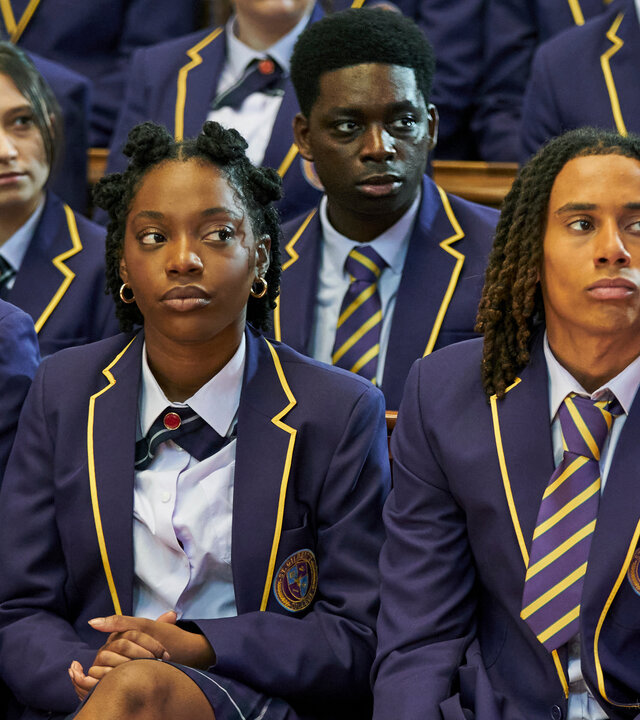 "Boarders - Welcome to St. Gilbert's- Piercings & Protests": Jaheim (Josh Tedeku), Leah (Jodie Campbell), Toby (Sekou Diaby), Omar (Myles Kamwendo) und Femi (Aruna Jalloh) sitzen mit Mitschülerinnen und Mitschülern (Komparsen) in der Aula. Alle tragen ihre Schuluniform und blicken ernst zur Seite.