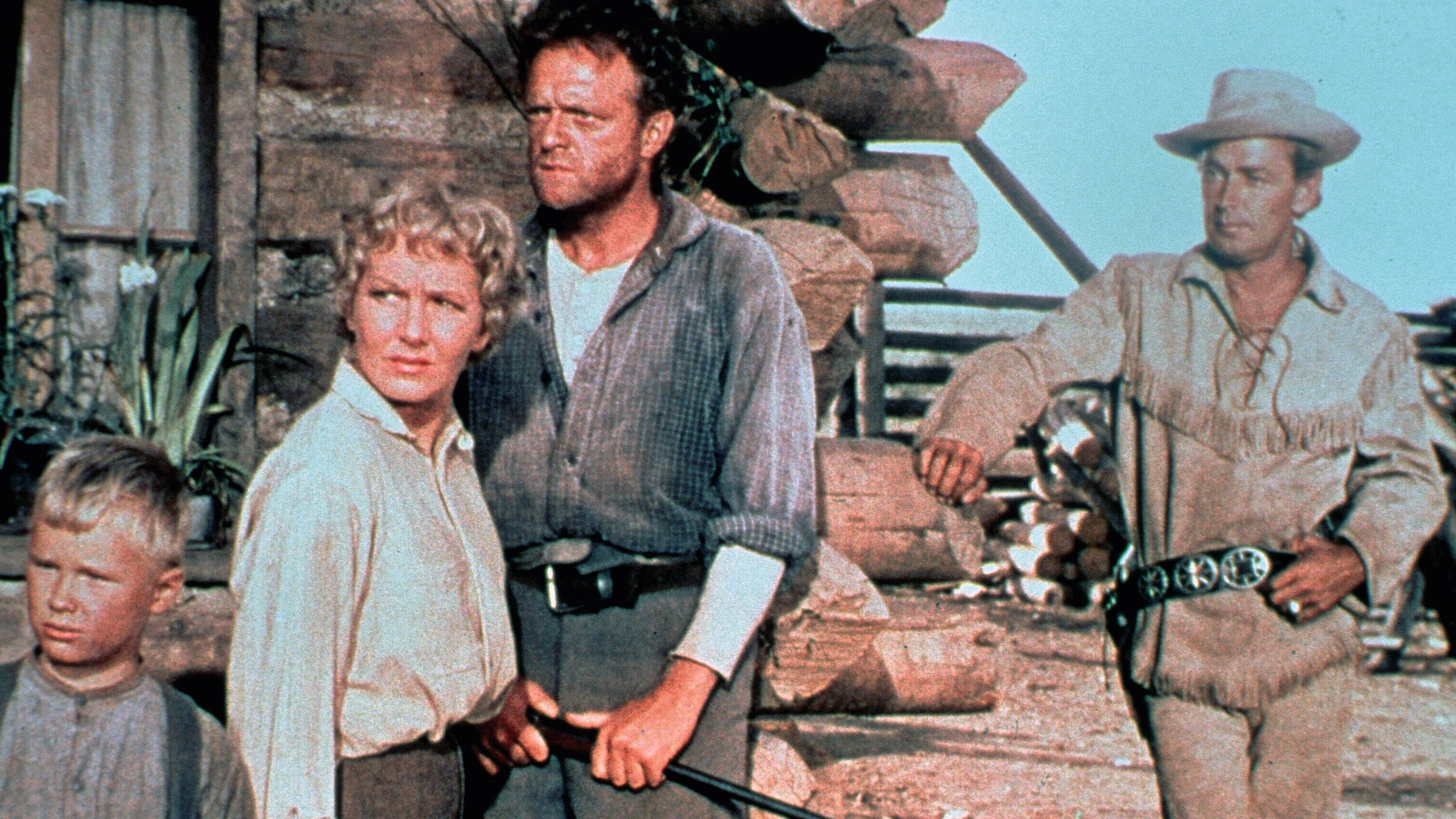 "Mein großer Freund Shane": An der Ecke einer Blockhütte steht ein kleiner Junge, eine Frau und ein Mann dicht beieinander. Sie schauen besorgt. Ein anderer Mann mit Hut und Pistolengurt steht hinter ihnen und beobachtet gelassen die Situation.