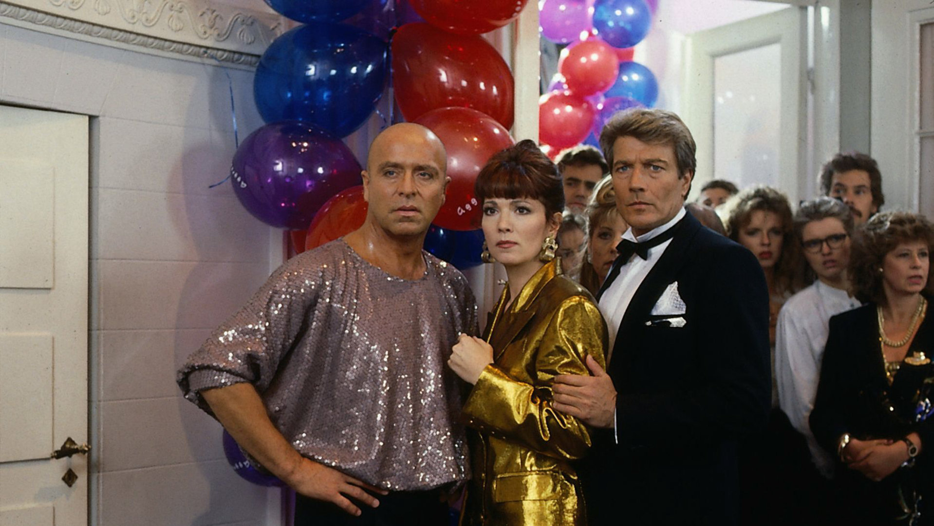 "Das Erbe der Guldenburgs - Das letzte Kapitel": Achim (Wilfried Baasner), Evelyn (Iris Berben) und Maurice Bernard (Amadeus August) stehen auf ihrer Modeparty und schauen ungläubig. Im Hintergrund hängen bunte Luftballons.