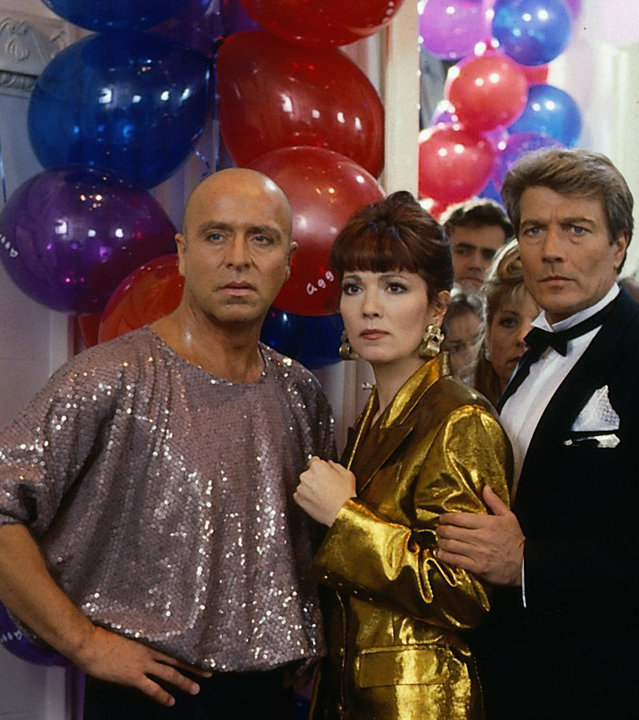 "Das Erbe der Guldenburgs - Das letzte Kapitel": Achim (Wilfried Baasner), Evelyn (Iris Berben) und Maurice Bernard (Amadeus August) stehen auf ihrer Modeparty und schauen ungläubig. Im Hintergrund hängen bunte Luftballons.