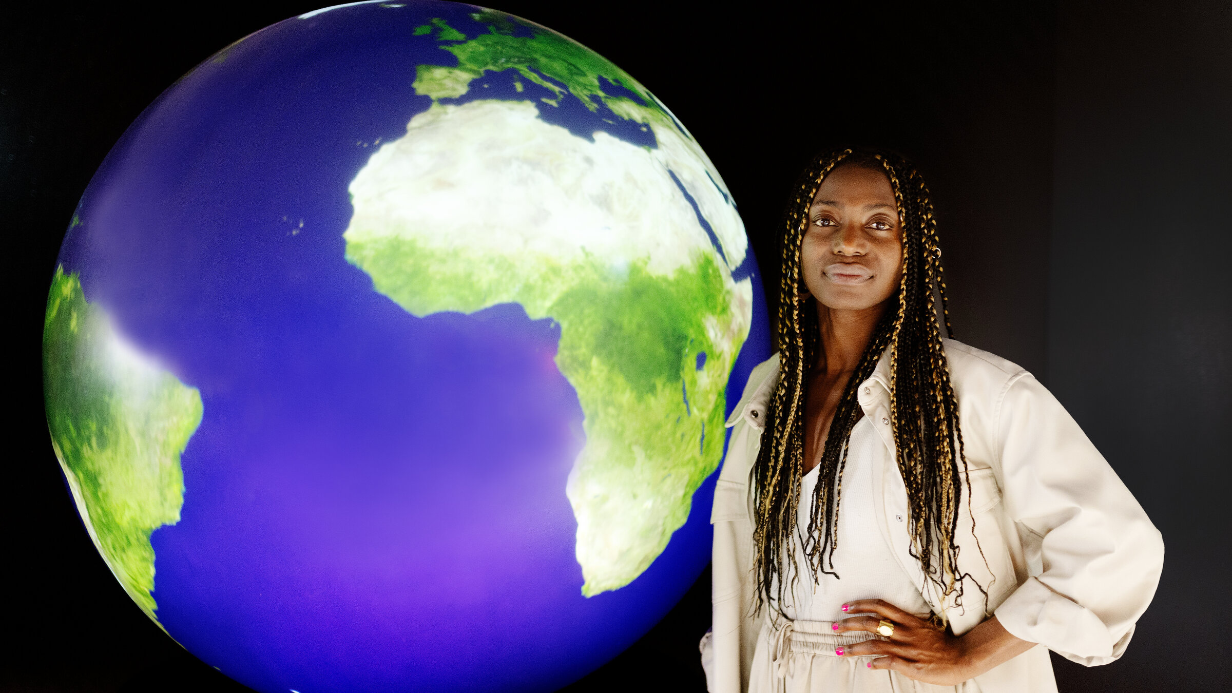 "Staatenlos - Kein Pass, keine Rechte": Christiana steht vor einem schwarzen Hintergrund, neben ihr steht ein beleuchteter Globus.