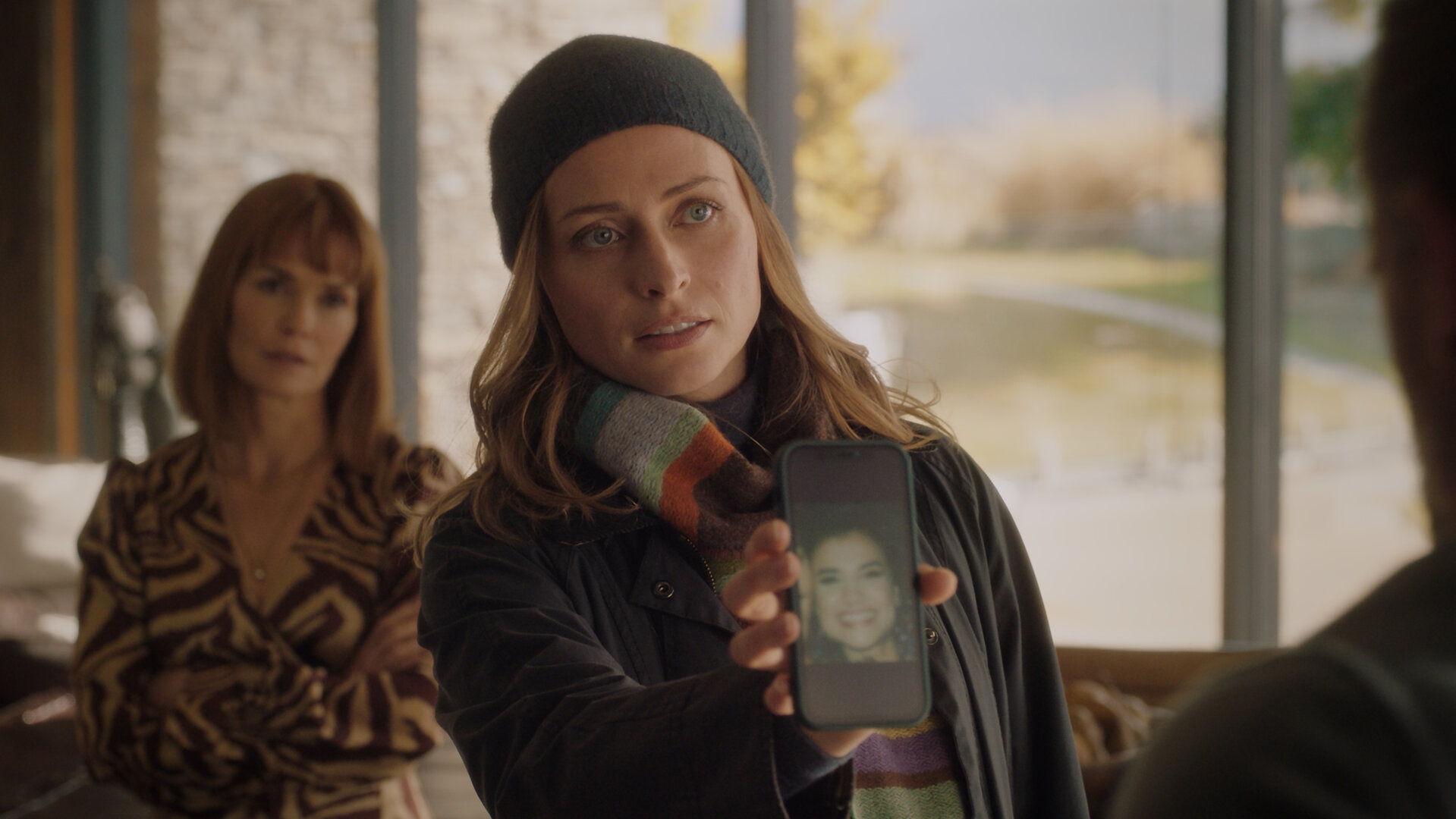 "Queenstown Murders – Zum Sterben schön - Am Abgrund": Detective Sergeant Anais Mallory (Chelsie Preston Crayford) zeigt Sebastian van Heusen (Mike Edward) ein Handy-Display mit dem Foto einer Frau. Er ist nur ansatzweise im Profil zu sehen. Aus dem Hintergrund beobachtet Marijke van Heusen (Nathalie Boltt) den Vorgang. Sie wirkt verärgert.