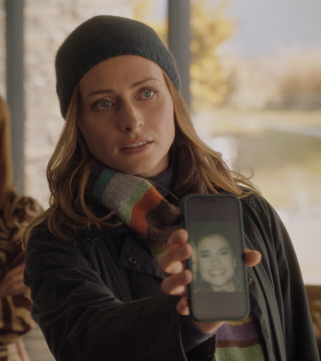 "Queenstown Murders – Zum Sterben schön - Am Abgrund": Detective Sergeant Anais Mallory (Chelsie Preston Crayford) zeigt Sebastian van Heusen (Mike Edward) ein Handy-Display mit dem Foto einer Frau. Er ist nur ansatzweise im Profil zu sehen. Aus dem Hintergrund beobachtet Marijke van Heusen (Nathalie Boltt) den Vorgang. Sie wirkt verärgert.