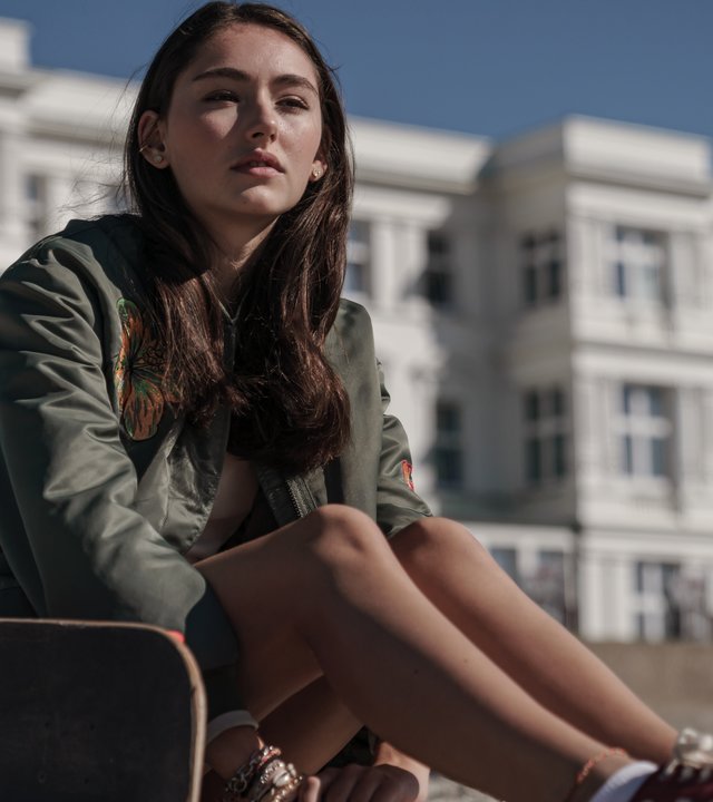 Sløborn: Evelin (Emily Kusche) sitzt am Strand und sieht nachdenklich nach vorne. Neben ihr liegt ihr Skateboard.