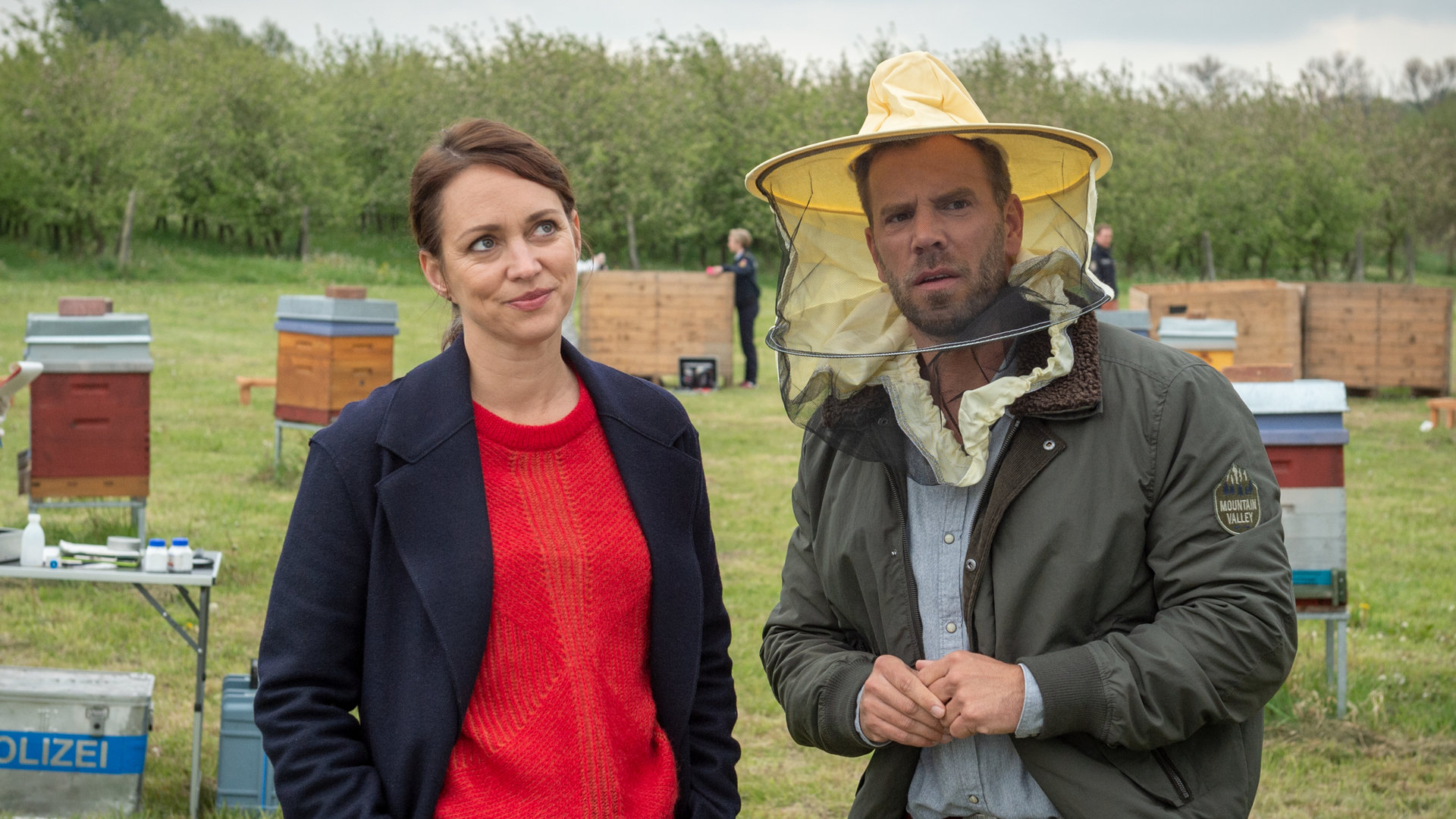 „SOKO Wismar – Für eine Handvoll Blüten“: Auf einer großen Wiese, die von Sträuchern im Hintergrund eingesäumt wird, stehen Karoline Joost (Nike Fuhrmann) und Lars Pöhlmann (Dominic Boeer). Kommissar Pöhlmann trägt einen auffälligen Imkerhelm, um sich vor den Bienen zu schützen. Sein Blick ist leicht panisch. Karoline Joost verdreht schmunzelnd die Augen.