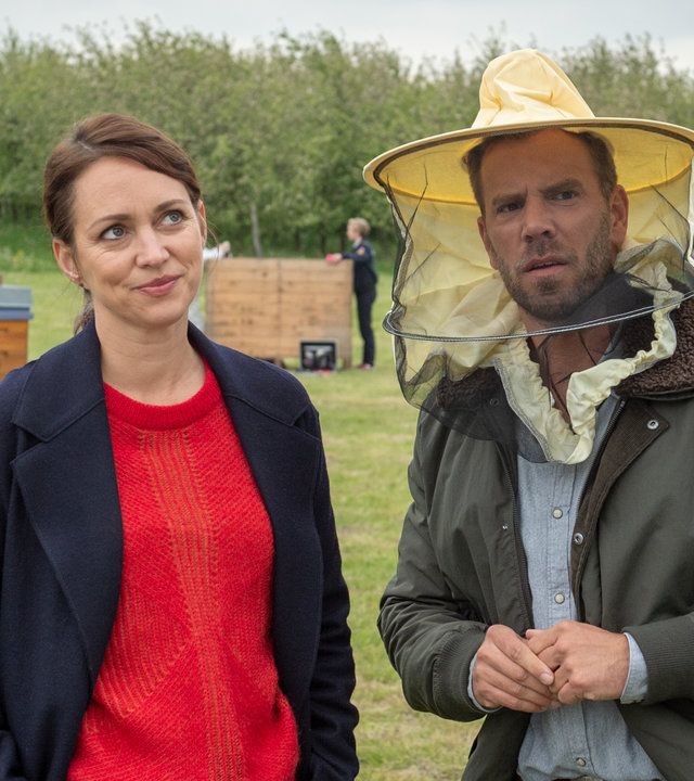 „SOKO Wismar – Für eine Handvoll Blüten“: Auf einer großen Wiese, die von Sträuchern im Hintergrund eingesäumt wird, stehen Karoline Joost (Nike Fuhrmann) und Lars Pöhlmann (Dominic Boeer). Kommissar Pöhlmann trägt einen auffälligen Imkerhelm, um sich vor den Bienen zu schützen. Sein Blick ist leicht panisch. Karoline Joost verdreht schmunzelnd die Augen.