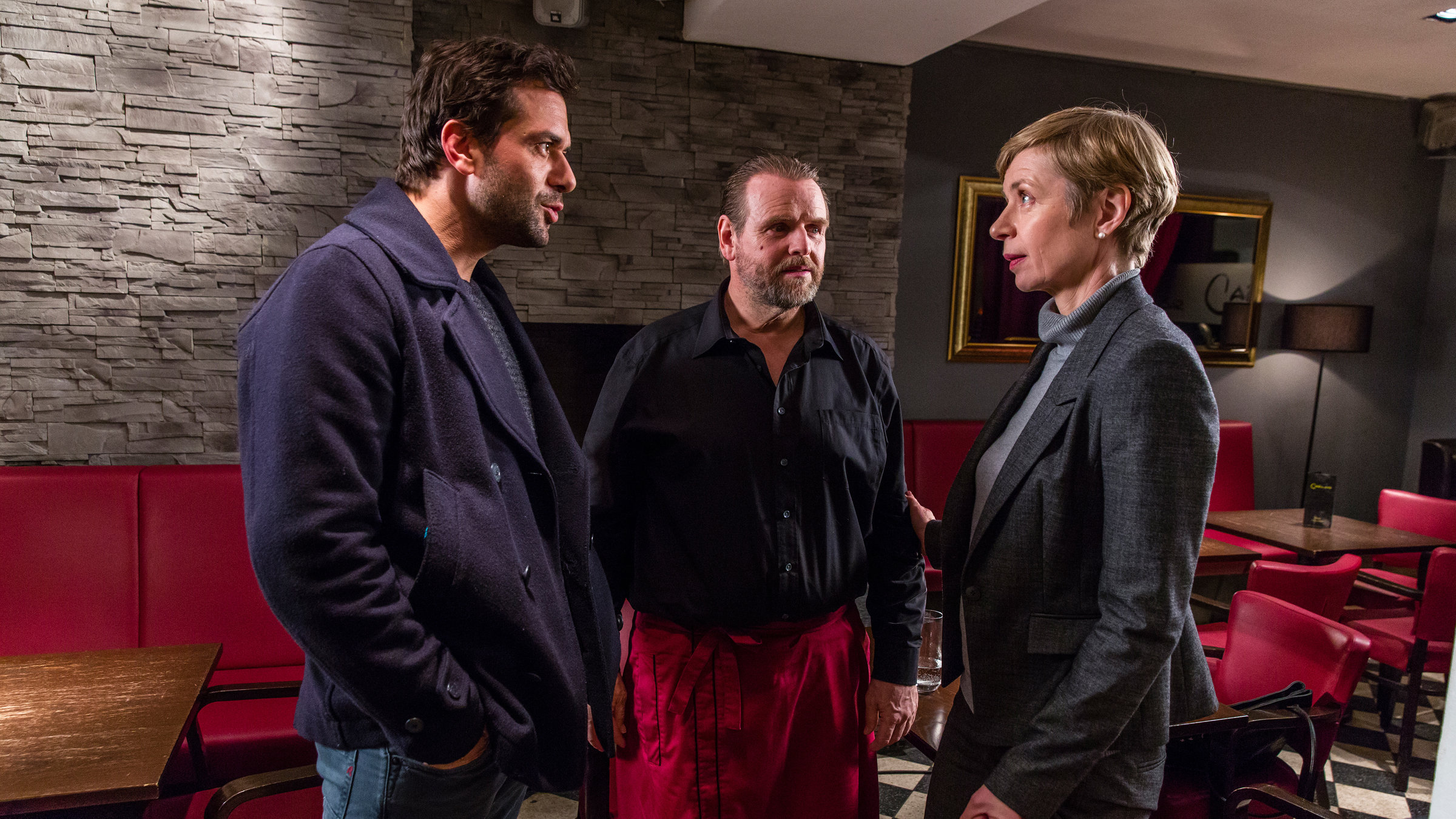 "Heldt - Carlos letzte Liebe": Heldt (Kai Schumann), Carlo (Felix Vörtler) und Anne Lerch (Anna Stieblich) stehen sich in Carlos Bar gegenüber und reden. Im Hintergrund sind Holztische und rote Stühle und Bänke zu sehen.