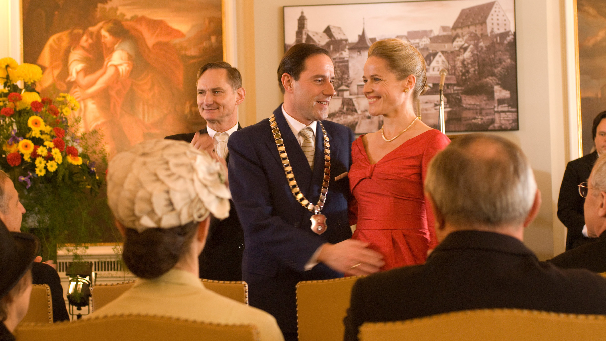 "Die Frau, die im Wald verschwand": In einem hell erleuchteten Festsaal steht ein Paar vor einem sitzenden Publikum. Der Mann, der Anzug und Krawatte trägt, hat eine große goldene Kette umgehängt und berührt mit der rechten Hand sanft den Bauch seiner Frau, die neben ihm steht in einem roten Kleid und mit hochgesteckten blonden Haaren. Beide lächeln. An der Wand im Hintergrund hängt das Foto einer Stadt und ein historisches Gemälde. Ein Mann neben dem Paar klatscht.