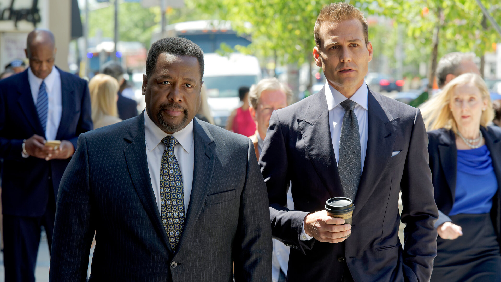 "Suits - Geschäftsführende Partner": Robert (Wendell Pierce, l.) und Harvey (Gabriel Mach) laufen auf einer Straße New Yorks nebeneinander. Harvey trägt in seiner rechten Hand einen Coffee-to-go. Beide Männer tragen elegante, dunkle Anzüge. Im Hintergrund sind weitere Personen zu erkennen.