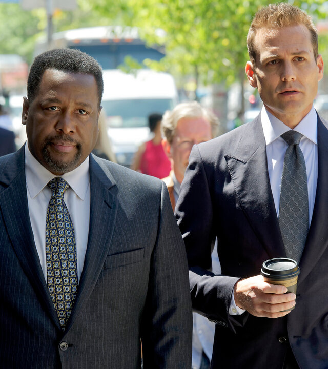 "Suits - Geschäftsführende Partner": Robert (Wendell Pierce, l.) und Harvey (Gabriel Mach) laufen auf einer Straße New Yorks nebeneinander. Harvey trägt in seiner rechten Hand einen Coffee-to-go. Beide Männer tragen elegante, dunkle Anzüge. Im Hintergrund sind weitere Personen zu erkennen.