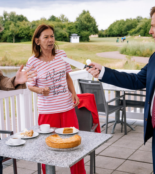 "SOKO Köln - Make Dünnwald Great Again": Tillmann Küppers (Martin Klempnow) steht Hauptkommissar Matti Wagner (Pierre Besson) auf einer Terrasse gegenüber und hat drohend ein großes Messer auf ihn gerichtet. Matti hält beschwichtigend seine linke Hand nach oben und sieht Tillmann Küppers aufgebracht an. Neben dem Kommissar steht Maria Küppers (Susanne Bredehöft), die ihren Sohn fassungslos ansieht. Zwischen den beiden Männern steht ein kleiner, grauer Marmortisch, auf dem sich ein Kuchen, zwei Teller mit Kuchenstücken und zwei Kaffeetassen befinden.