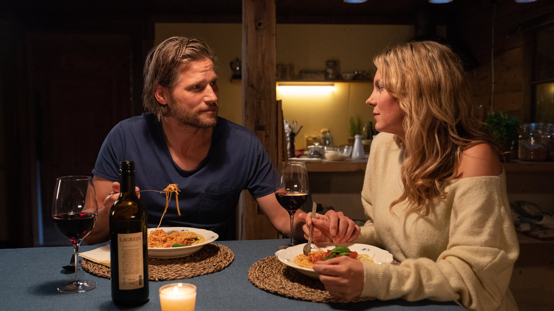 "Die Bergretter: Ausgesetzt (2)": Katharina (Luise Bähr) und Markus (Sebastian Ströbel) sitzen bei gedämpften Licht am Küchentisch. Katharina blickt Markus traurig an, der aufmunternd seine Hand auf ihre gelegt hat. Vor ihnen stehen Teller mit Spaghetti und Rotweingläser.