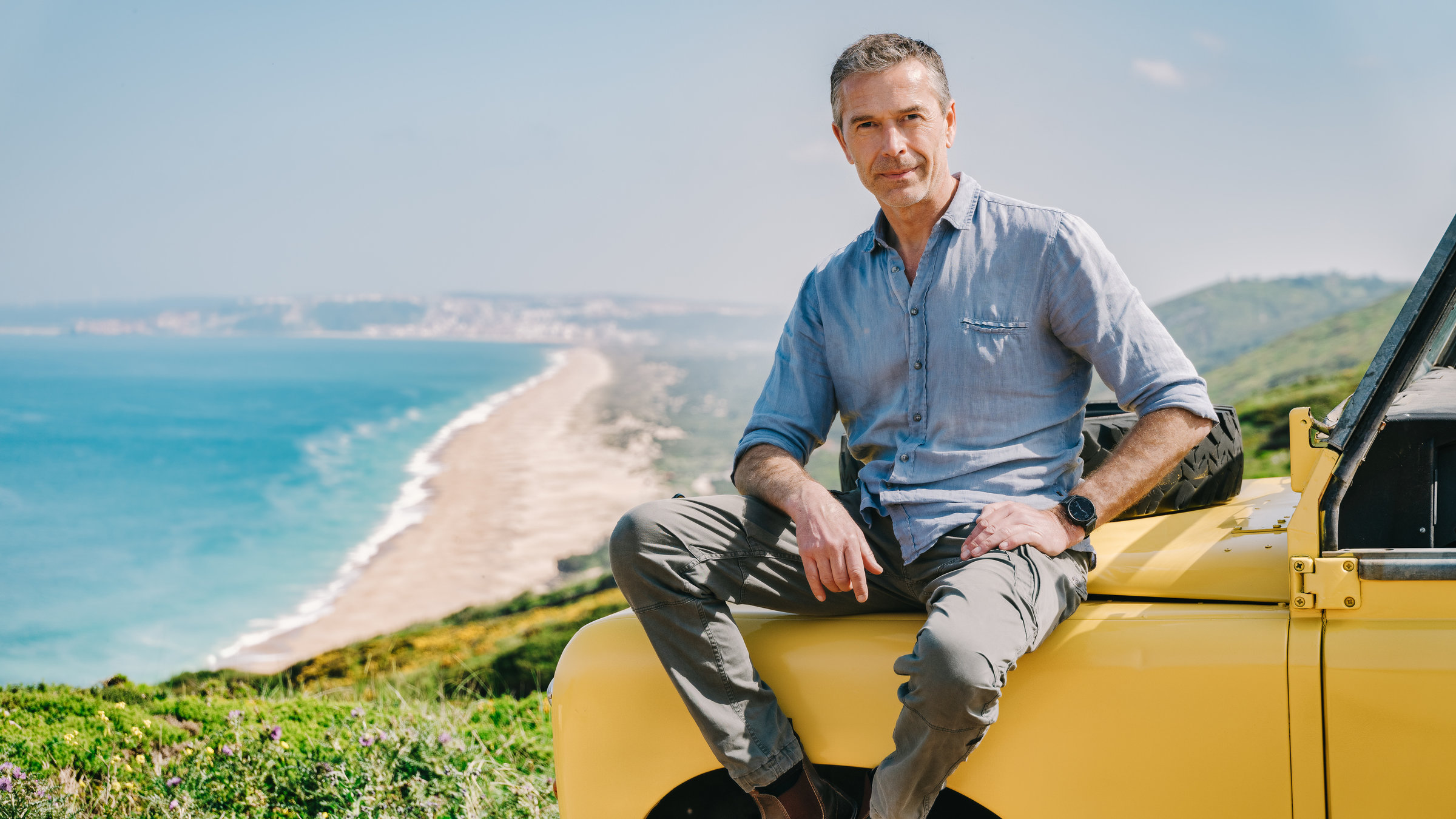 "Terra X: Rätselhafte Phänomene - Monsterwellen und Fluggiganten": Der Moderator Dirk Steffens sitz auf der Motorhaube eines gelben Jeeps, der auf einer hohen Klippe steht. Im Hintergrund ist ein heller Strand und das blaue Meer zu sehen.