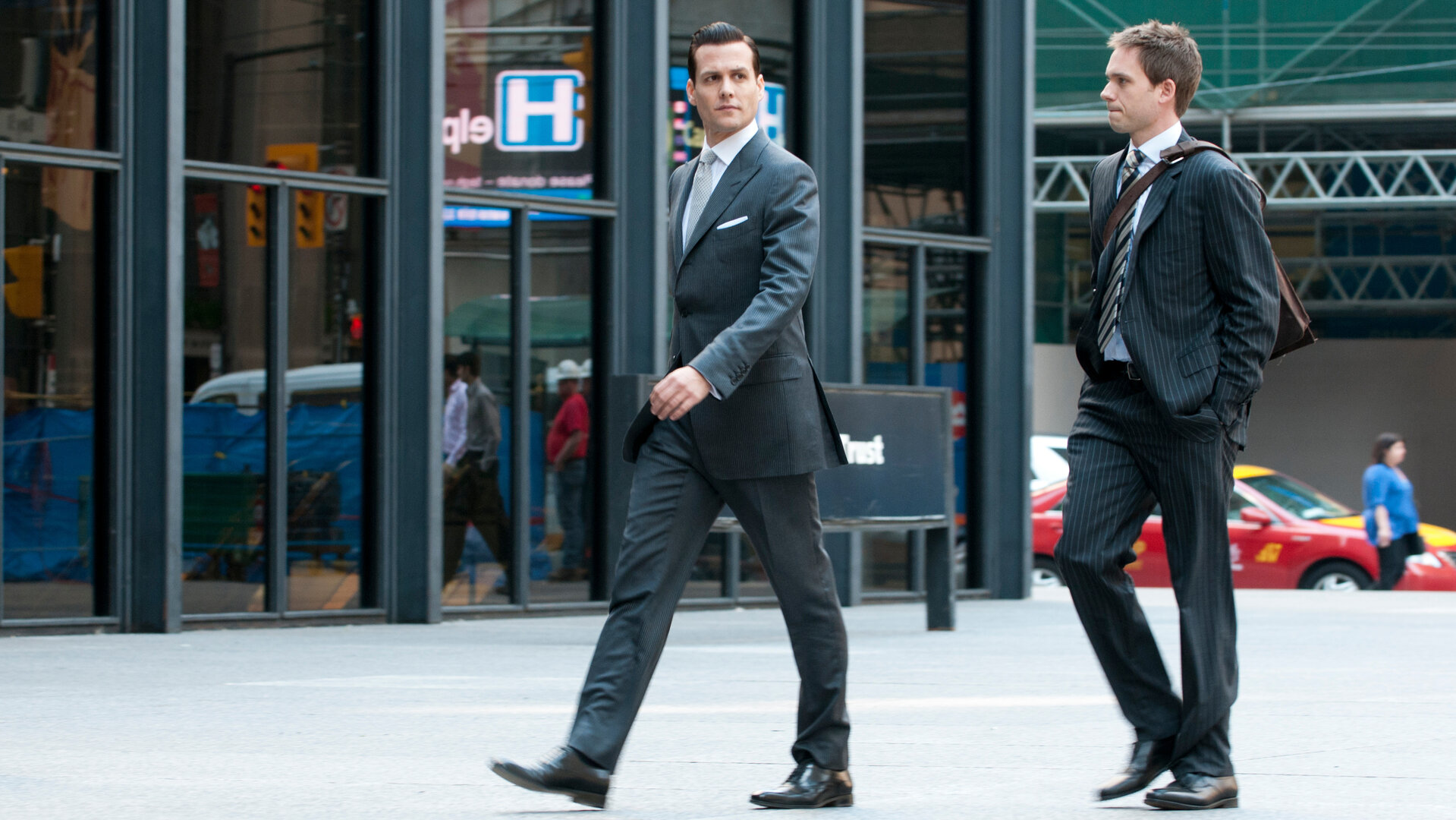 "Suits - Schmutzige kleine Geheimnisse": Harvey (Gabriel Macht) und Mike (Patrick J. Adams) gehen nebeneinander an einer gläsernen Häuserfront entlang. Beide tragen dunkle Anzüge, Mike hat die Hände in den Hosentaschen, Harvey blickt entschlossen zur Seite.