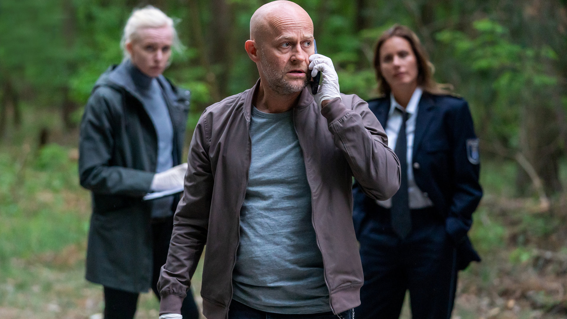 "Jenseits der Spree - Zwischen den Welten": Robert Heffler (Jürgen Vogel) telefoniert in einem Waldstück mit seinem Mobiltelefon. Katharina Koblinski (Elisabeth Baulitz) und Veronika Schäfer (Anne Müller) beobachten ihn aus dem Hintergrund.