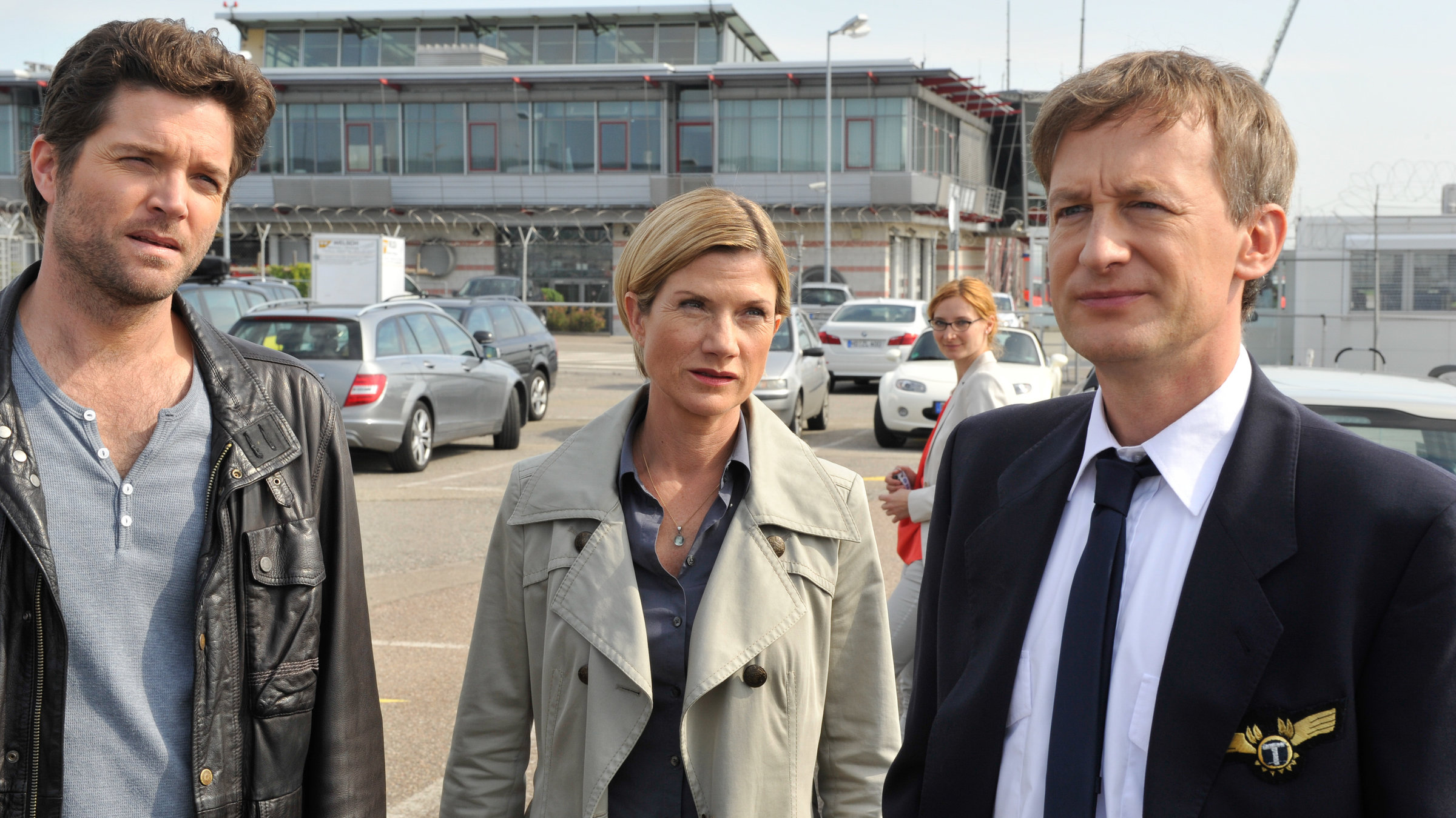 "SOKO Stuttgart - Abflug": Jo Stoll (Peter Ketnath) und Martina Seiffert (Astrid M. Fünderich) befragen den Piloten Kai-Uwe Maassen (Jan Henrik Stahlberg) auf einem Parkplatz. Seine Frau Bea (Komparsin) steht etwas abseits hinter ihnen.