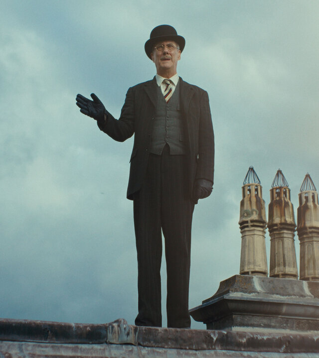 "This Is Not a Murder Mystery - Die Phantomlady": DCI John Thistlethwaite (Stephen Tompkinson) steht auf einem Dachfirst. Neben von ihm ragen mehrere kleine Schornsteine in den Himmel. Er trägt einen Anzug mit Krawatte, Handschuhe und eine Melone. Mit der rechten Hand deutet er zur Seite.