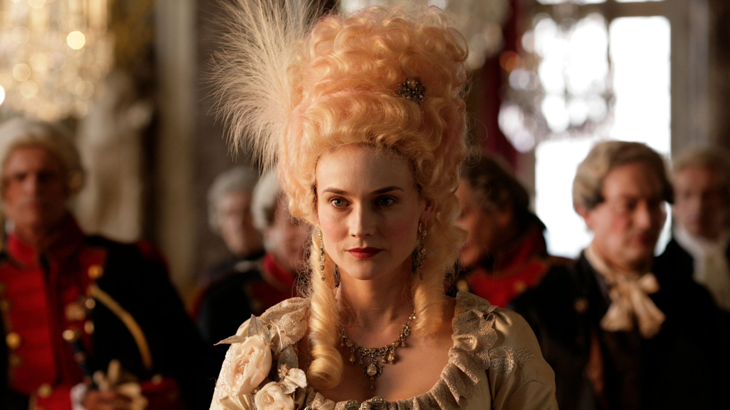 „Leb wohl, meine Königin!“: Großaufnahme – Mit wunderbarer, orange-rot schillernder Steckfrisur, langen Locken an beiden Seiten des Gesichts, und erlesenem Kleid mit Dekolleté und Perlenkette, tritt Marie-Antoinette (Diane Kruger) in einem Saal am Hof auf. Im Hintergrund sind unscharf Männer in entsprechender Kleidung zu sehen.