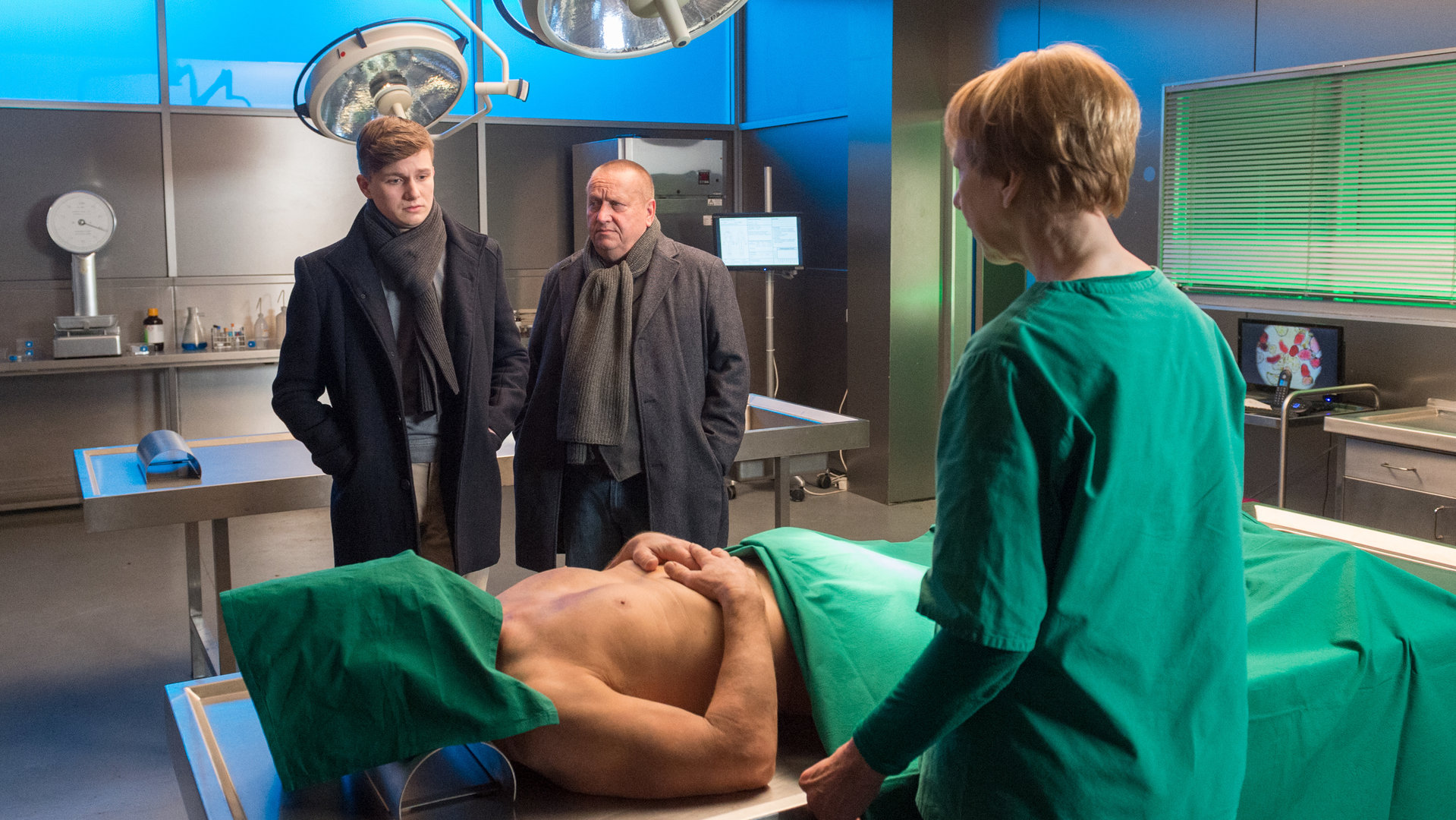 "SOKO Wismar – Lügen können tödlich sein": Die am Kopf und Unterleib abgedeckte Leiche von Karsten Walbeck (Hans Heller) liegt in der Rechtsmedizin auf dem Seziertisch. Bei ihr stehen Dr. Helene Sturbeck (Katharina Blaschke), Jan Reuter (Udo Kroschwald) und der Stiefsohn des Toten Niklas Fellner (Leon Blaschke). Niklas blickt traurig zu seinem toten Stiefvater herab.