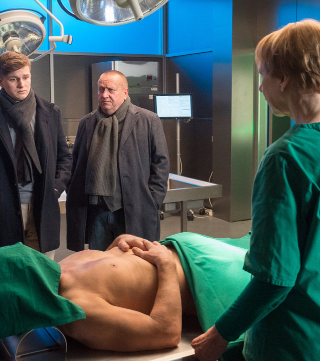 "SOKO Wismar – Lügen können tödlich sein": Die am Kopf und Unterleib abgedeckte Leiche von Karsten Walbeck (Hans Heller) liegt in der Rechtsmedizin auf dem Seziertisch. Bei ihr stehen Dr. Helene Sturbeck (Katharina Blaschke), Jan Reuter (Udo Kroschwald) und der Stiefsohn des Toten Niklas Fellner (Leon Blaschke). Niklas blickt traurig zu seinem toten Stiefvater herab.