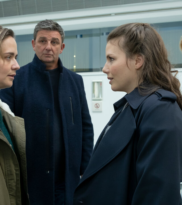 "Der Bergdoktor: Der Weg zurück": Im Krankenhaus: Katharina (Sarina Radomski) und Melanie Schober (Leonie Rainer) stehen sich gegenüber und schauen sich gebannt in die Augen. Im Hintergrund stehen Dr. Martin Gruber (Hans Sigl) und Dr. Angelika Rüdiger (Annika Ernst) und beobachten mit besorgten Blicken die beiden.