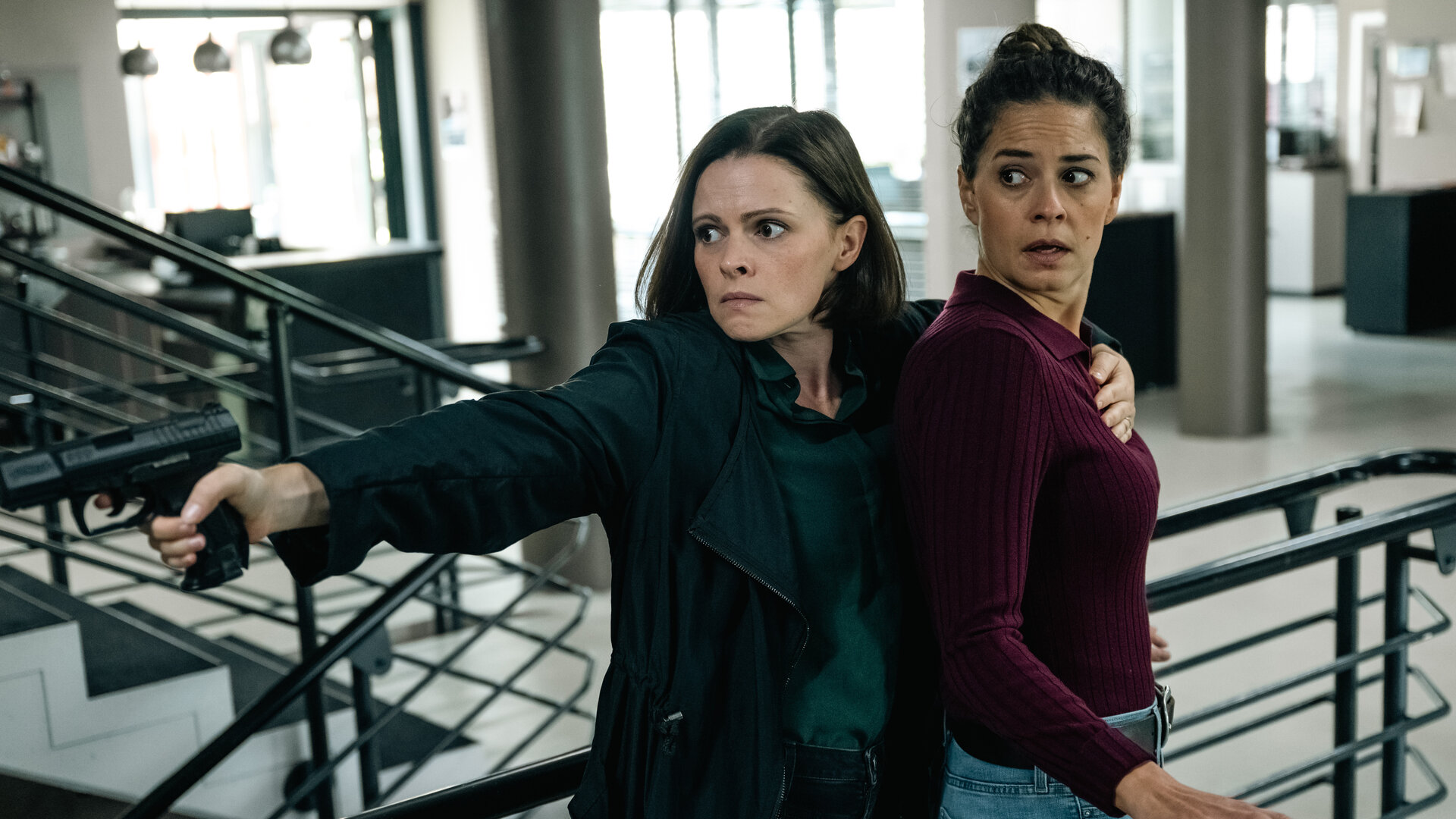 "SOKO Köln - Doppeltes Kaliber": Birgit Wilkens (Jennifer Ulrich) steht hinter Lilly Funke (Tatjana Kästel) im Kommissariat und legt ihr von hinten einen Arm um den Körper und eine Hand auf die Brust, um sie festzuhalten. In der anderen Hand hält sie eine Pistole, die sie mit angespanntem Blick zur Seite richtet. Lilly sieht ängstlich in die gleiche Richtung.