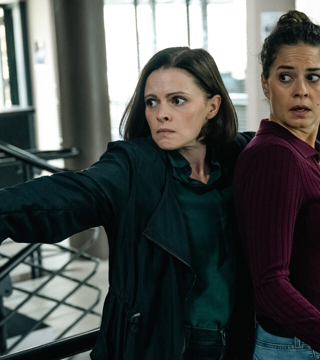 "SOKO Köln - Doppeltes Kaliber": Birgit Wilkens (Jennifer Ulrich) steht hinter Lilly Funke (Tatjana Kästel) im Kommissariat und legt ihr von hinten einen Arm um den Körper und eine Hand auf die Brust, um sie festzuhalten. In der anderen Hand hält sie eine Pistole, die sie mit angespanntem Blick zur Seite richtet. Lilly sieht ängstlich in die gleiche Richtung.