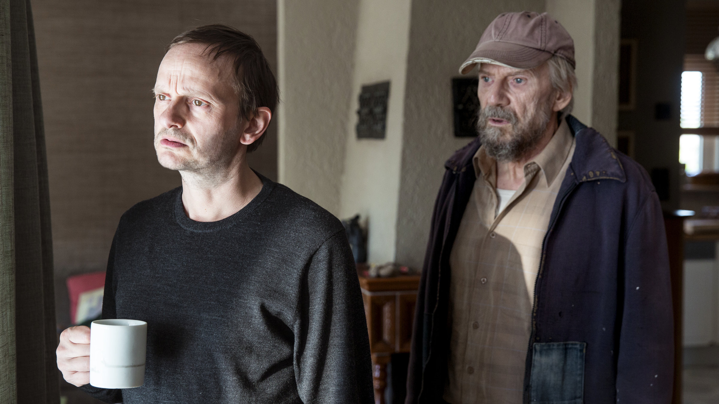 "Der Andere - Eine Familiengeschichte": Stefan (Milan Peschel) steht gedankenverloren mit einer Tasse in der Hand am Fenster. Im Hintergrund steht sein Vater Willi (Jesper Christensen).